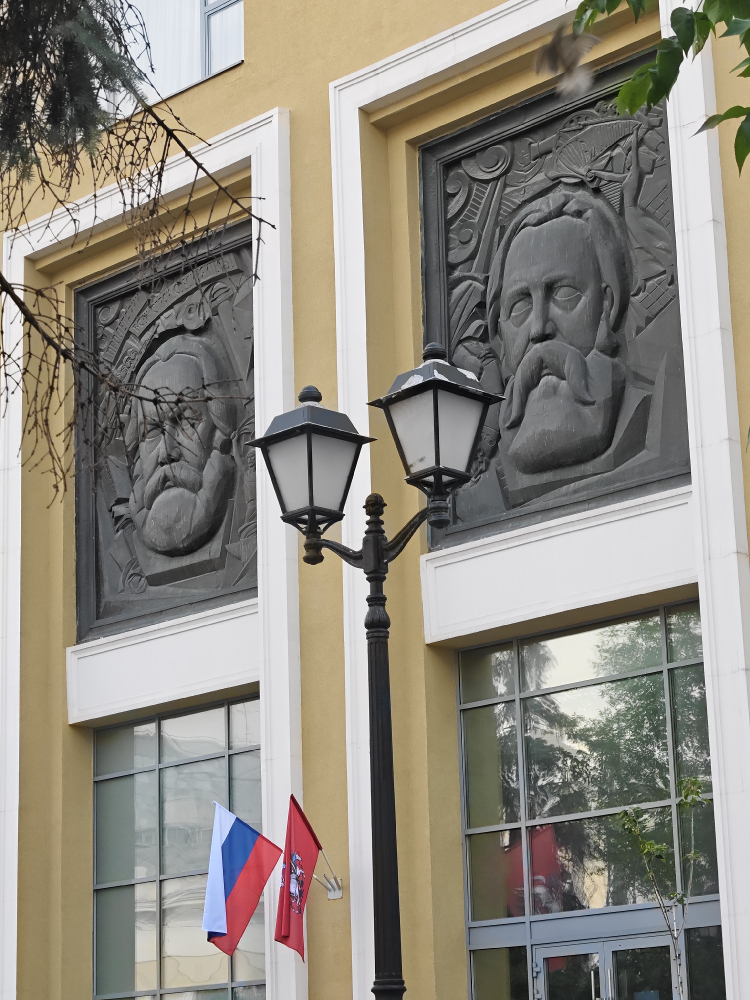 The image shows a detail of the facade of a building in Moscow, Russia. The building is painted a light yellow color. Two large, dark-colored bas-reliefs depicting male portraits are mounted on the facade, framed by white architectural elements. One relief is partially obscured by tree branches in the foreground. A black street lamp with a white glass enclosure stands in front of the bas-reliefs. Below the reliefs, large windows are visible, with reflections of trees and the sky. Two flags are displayed on the left side of the window: a Russian flag and a red flag with a coat of arms. The scene appears to be a daytime exterior shot.