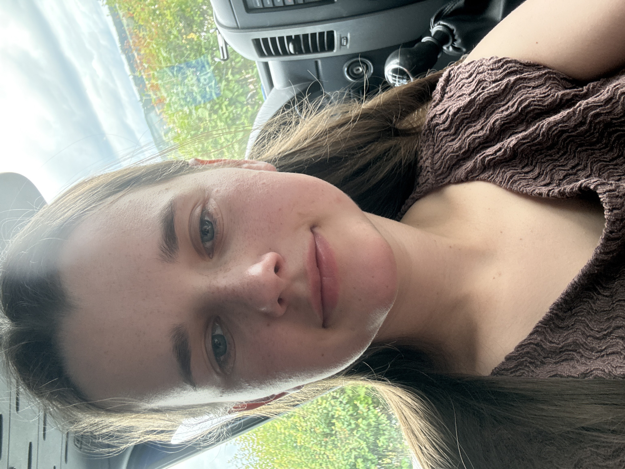 A young woman with light brown hair and blue eyes, wearing a brown textured top, is taking a selfie inside a vehicle. The background shows greenery and a cloudy sky, suggesting an outdoor setting. The vehicle's interior, including a gear shift and part of the dashboard, is visible on the right side of the image.