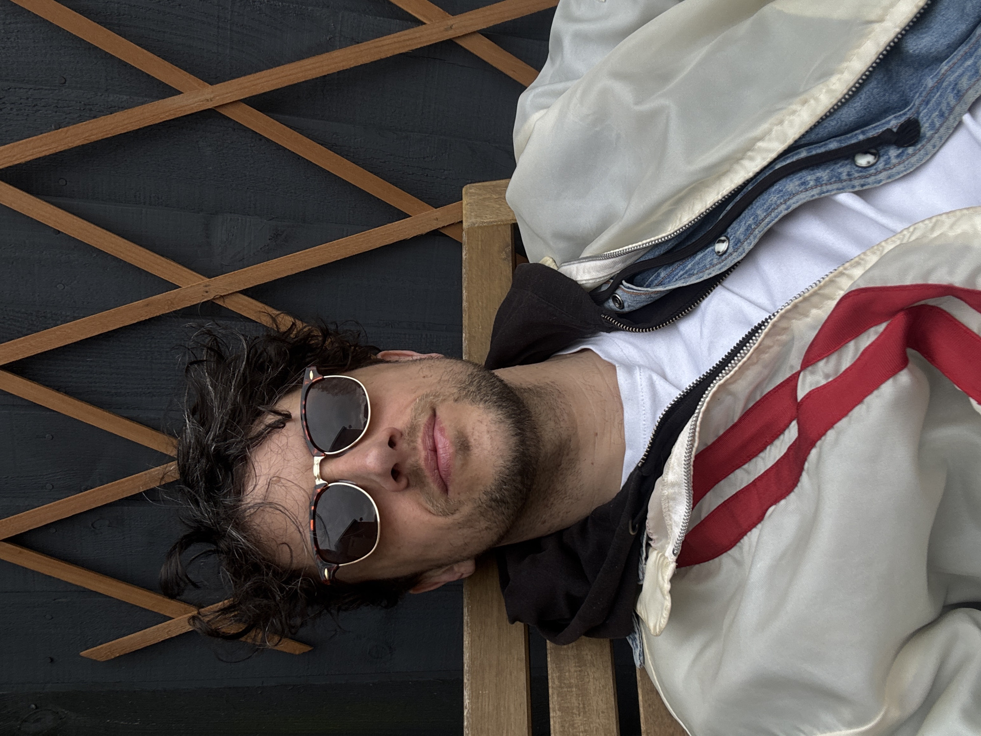 A man lies on a wooden bench against a dark wooden fence with a lattice overlay. He wears sunglasses and is dressed in a white t-shirt, a black hoodie, a denim jacket, and a white jacket with red stripes. His head rests on the bench with his hair spread out against the fence. The scene appears to be outdoors. No location data or reference to South Kesteven, United Kingdom, can be confirmed from the image itself.