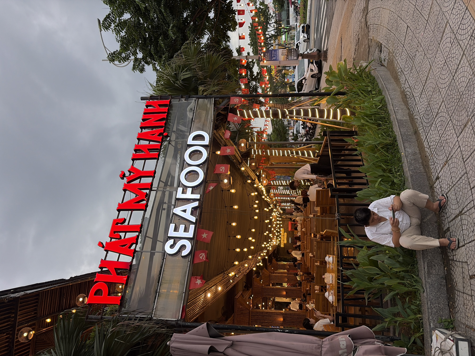 The image captures an outdoor dining area of a seafood restaurant named "PHAT MY HANH SEAFOOD" in Da Nang, Vietnam. The restaurant features a rustic aesthetic with bamboo accents and is illuminated by numerous string lights. Small red flags with yellow stars, reminiscent of national flags, are hung along the facade. A man is seated on a curb in the foreground, engrossed in his mobile phone. In the background, patrons are dining at wooden tables, and a street with vehicles is visible, indicating an urban setting. The sky is overcast, suggesting a cloudy day or evening.