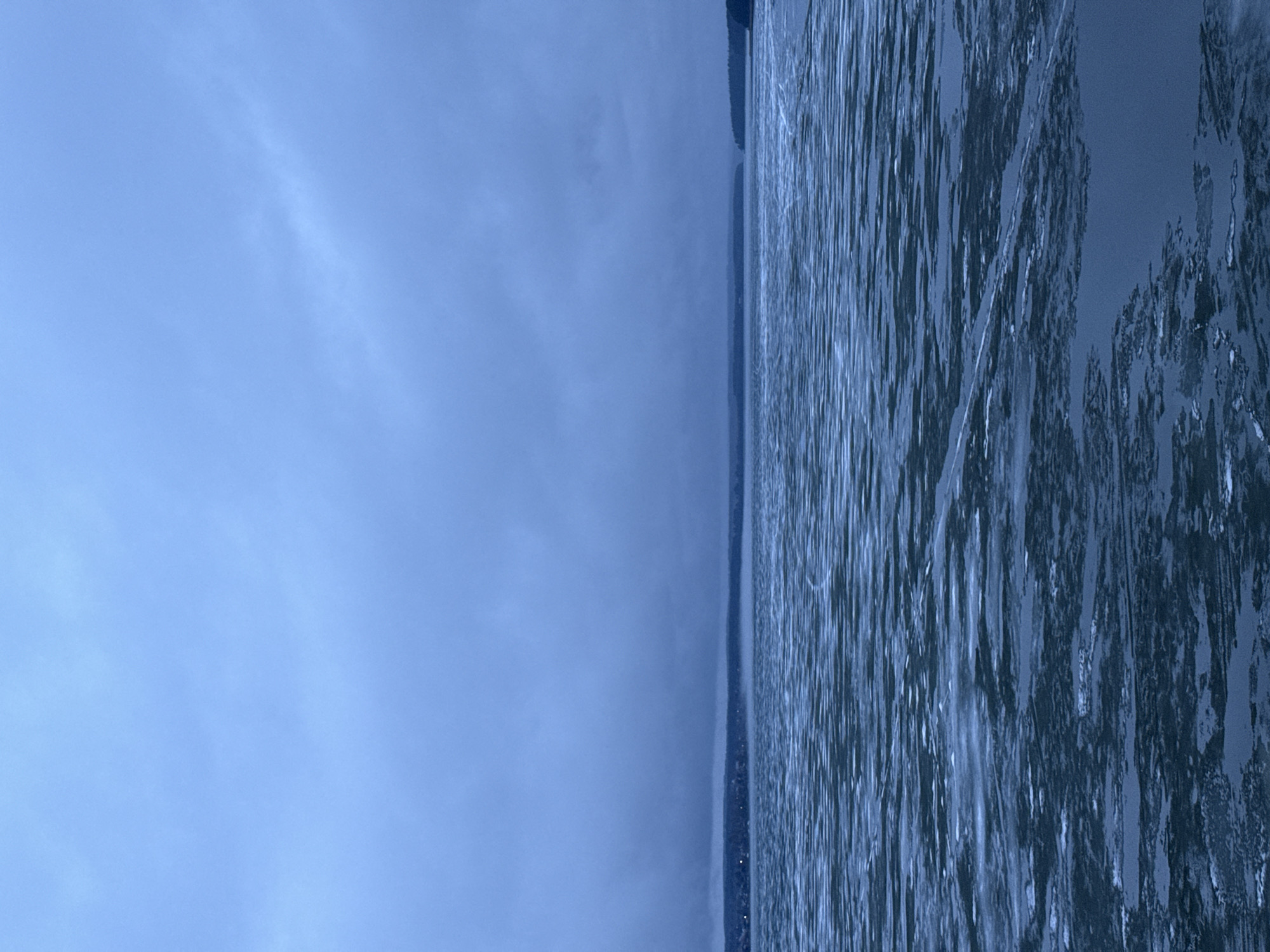 The image shows a body of water, possibly a lake or river, partially covered with ice, next to the city of Votkinsk, Russia. The sky is overcast and blue-toned. A strip of land is visible on the horizon between the sky and the water. The ice on the water appears to be thin and broken in places. There are no people or other subjects visible in the image. The image is rotated 90 degrees clockwise.