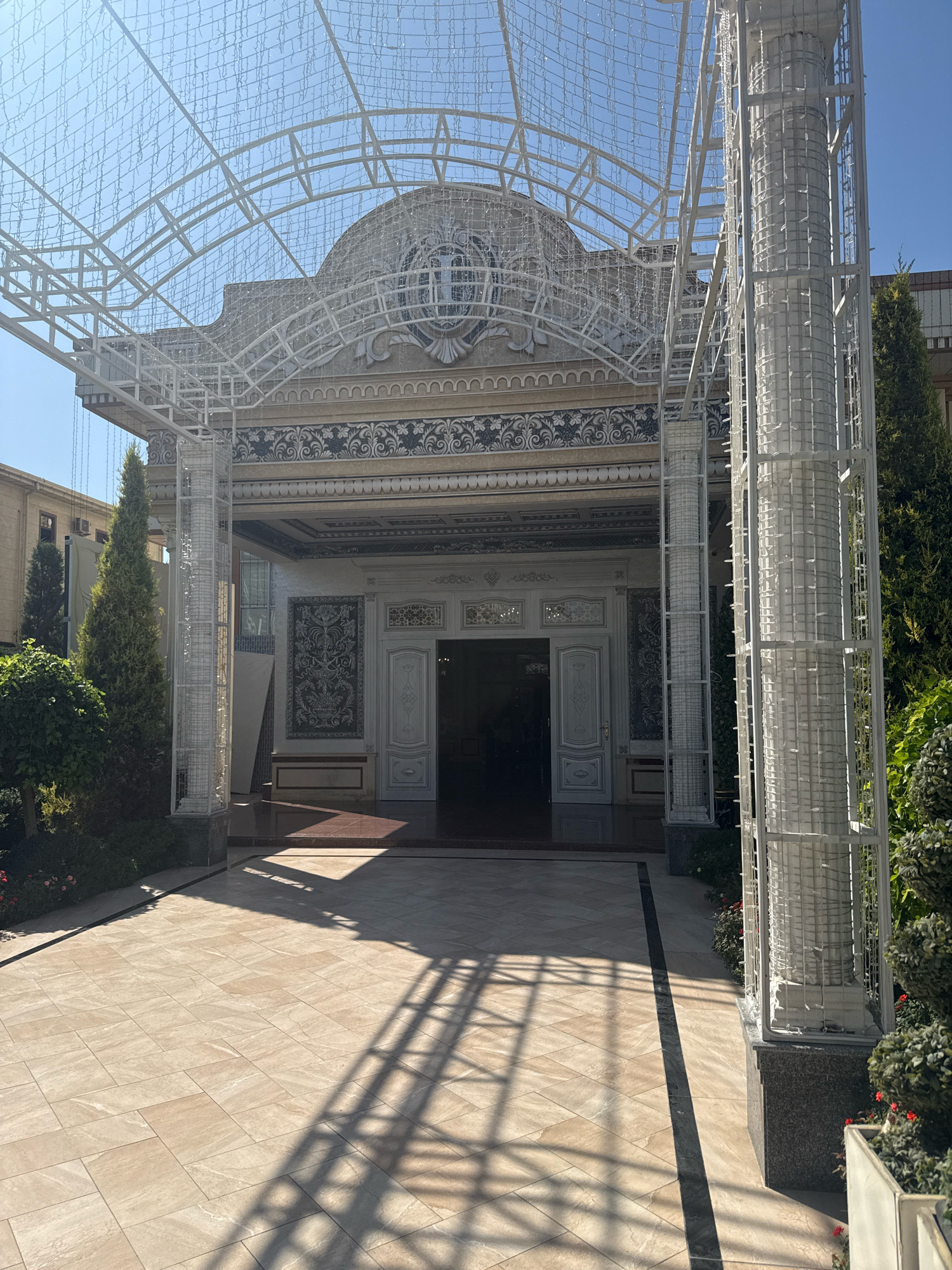 The image shows the exterior of a building with an ornate entrance, likely a wedding palace or similar venue, located in Darkhan, Uzbekistan. A white, elaborate canopy structure, possibly decorated with lights, extends over a tiled walkway leading to the entrance. The building itself features decorative patterns and a large, arched facade. Tall, slender trees flank the entrance. The sun is casting long shadows across the tiled ground. No people are visible in the image.