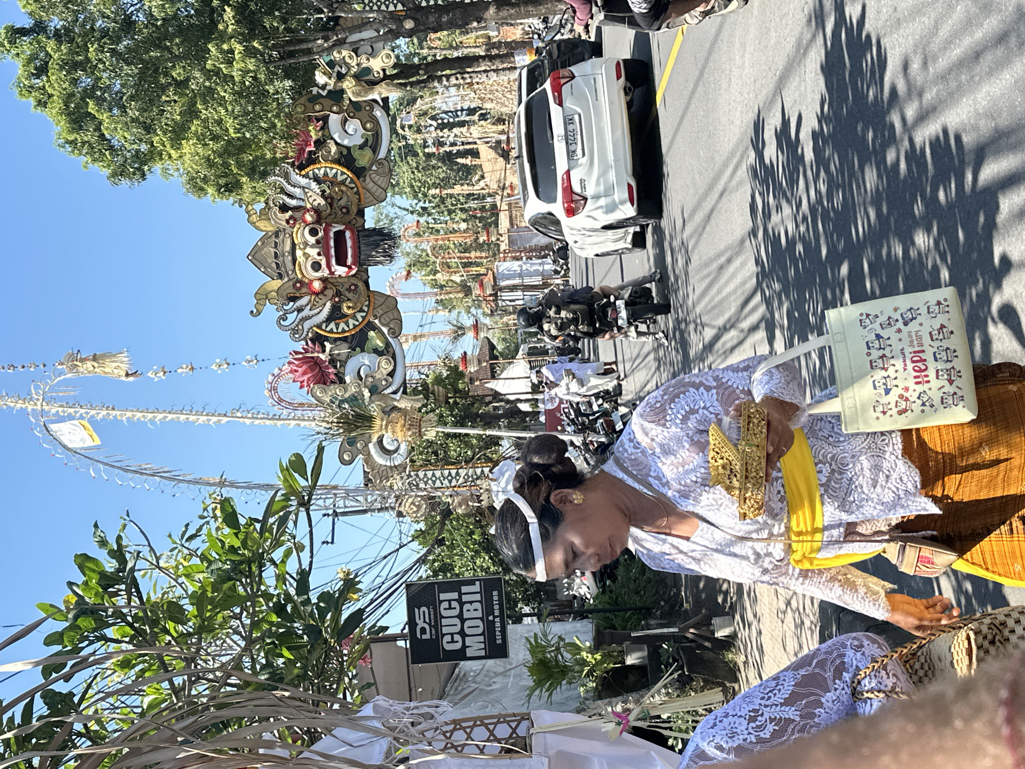In Sukawati, Indonesia, on a sunlit street, a woman wearing traditional Balinese attire, including a white lace top and a yellow sash, carries a golden offering tray and a tote bag. She stands on the right side of the road, amidst a procession or festival. Behind her, a white SUV with a license plate ending in "XK" is visible, along with other vehicles and motorcycles. Overhead, a decorative archway, possibly part of a temple or ceremonial entrance, features a prominent dragon-like face with red and gold accents. Trees line the street, casting dappled shadows on the asphalt. The scene suggests a cultural event taking place in a populated urban area.