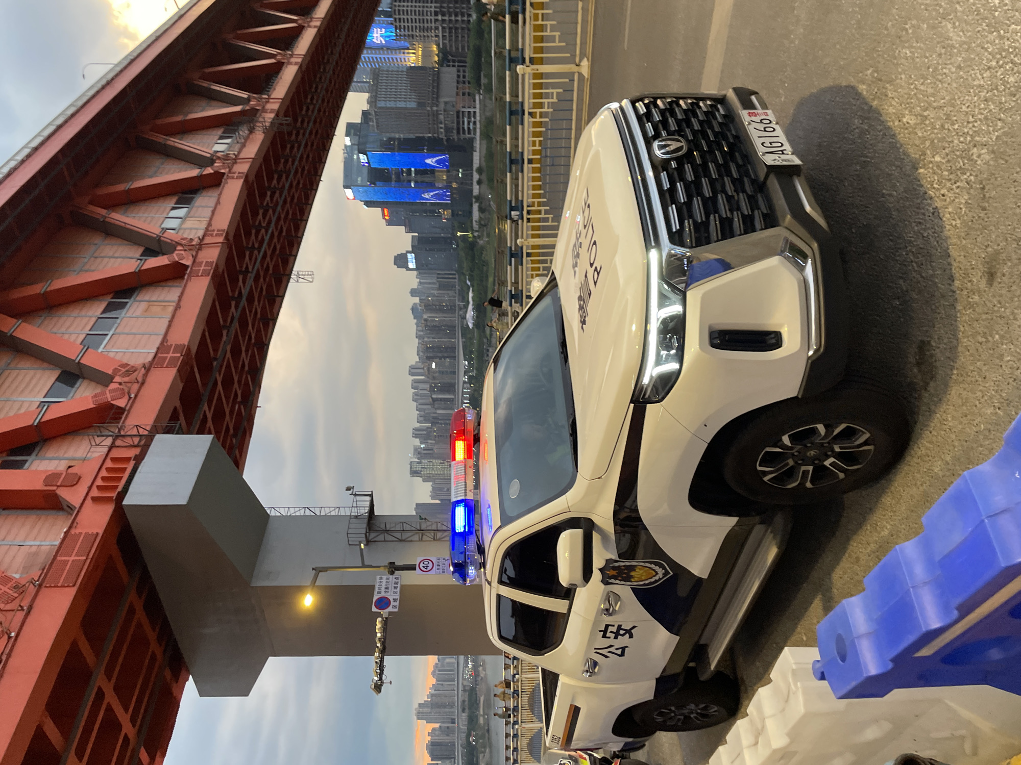 A white police pickup truck is parked on a bridge overlooking a city skyline. The truck has blue and red flashing lights on its roof and the word "Police" written on its hood and side. The truck is positioned in the foreground, with a large red bridge structure overhead. In the background, a dense urban landscape with numerous buildings, some illuminated with neon lights, stretches along a river. Several people are visible on the bridge walkway in the mid-ground, appearing to be walking or standing. The sky is overcast with hints of sunset colors. The scene appears to be set in the Yuzhong District of Chongqing, China, given the architecture and the presence of a major bridge over a river.