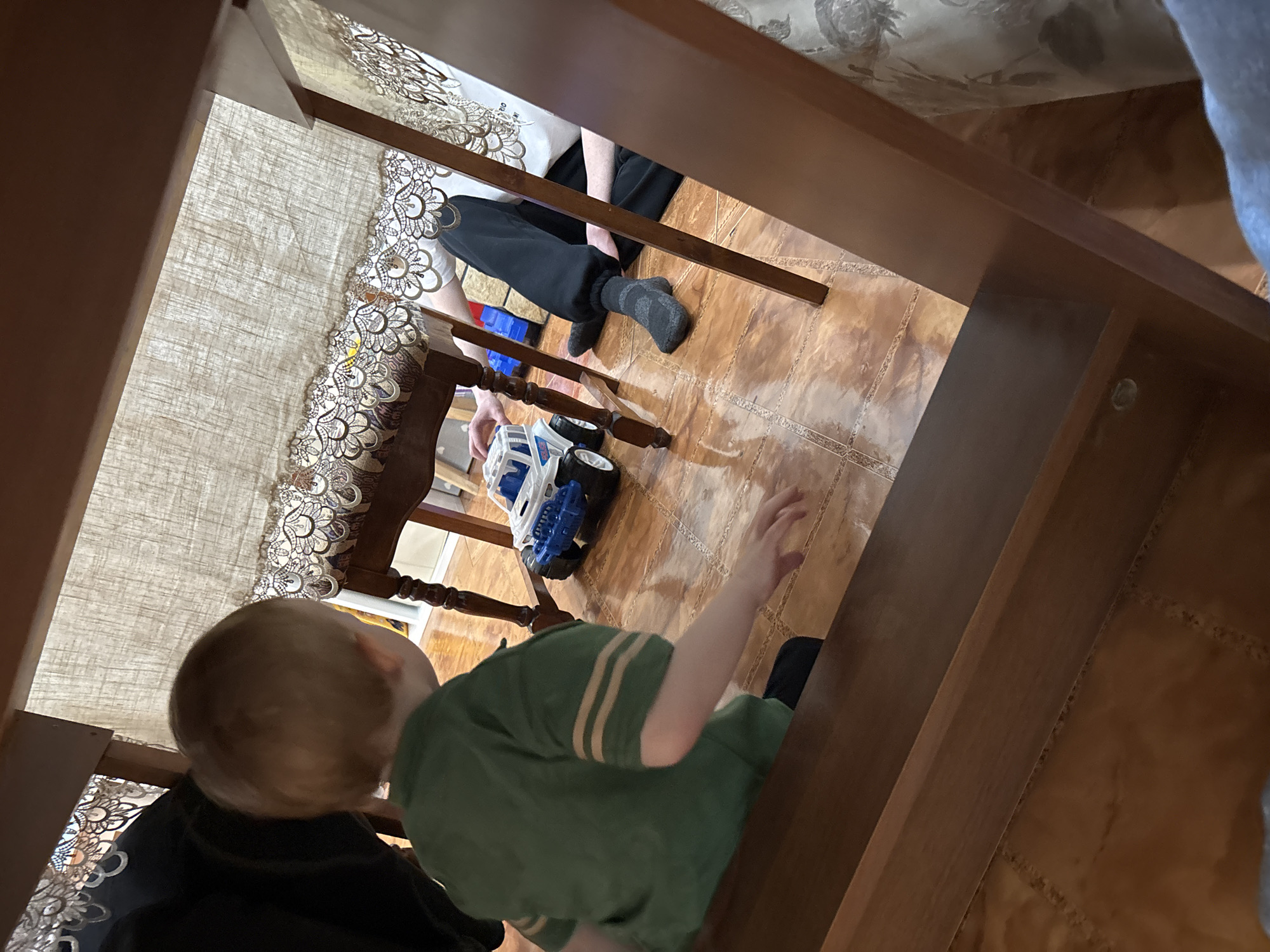 The image, shot from an unusual, upward-angled perspective, depicts an interior scene. A young child with blond hair, wearing a green shirt with light-colored stripes, is present. A person wearing black pants and dark socks is also visible in the background, near a toy truck. The scene takes place on a tiled floor. The setting includes elements of furniture, like a table or a chair, and a tablecloth with a lace trim. The wallpaper has a floral pattern. While the location is given as Ферма, Russia, nothing in the image specifically confirms this.