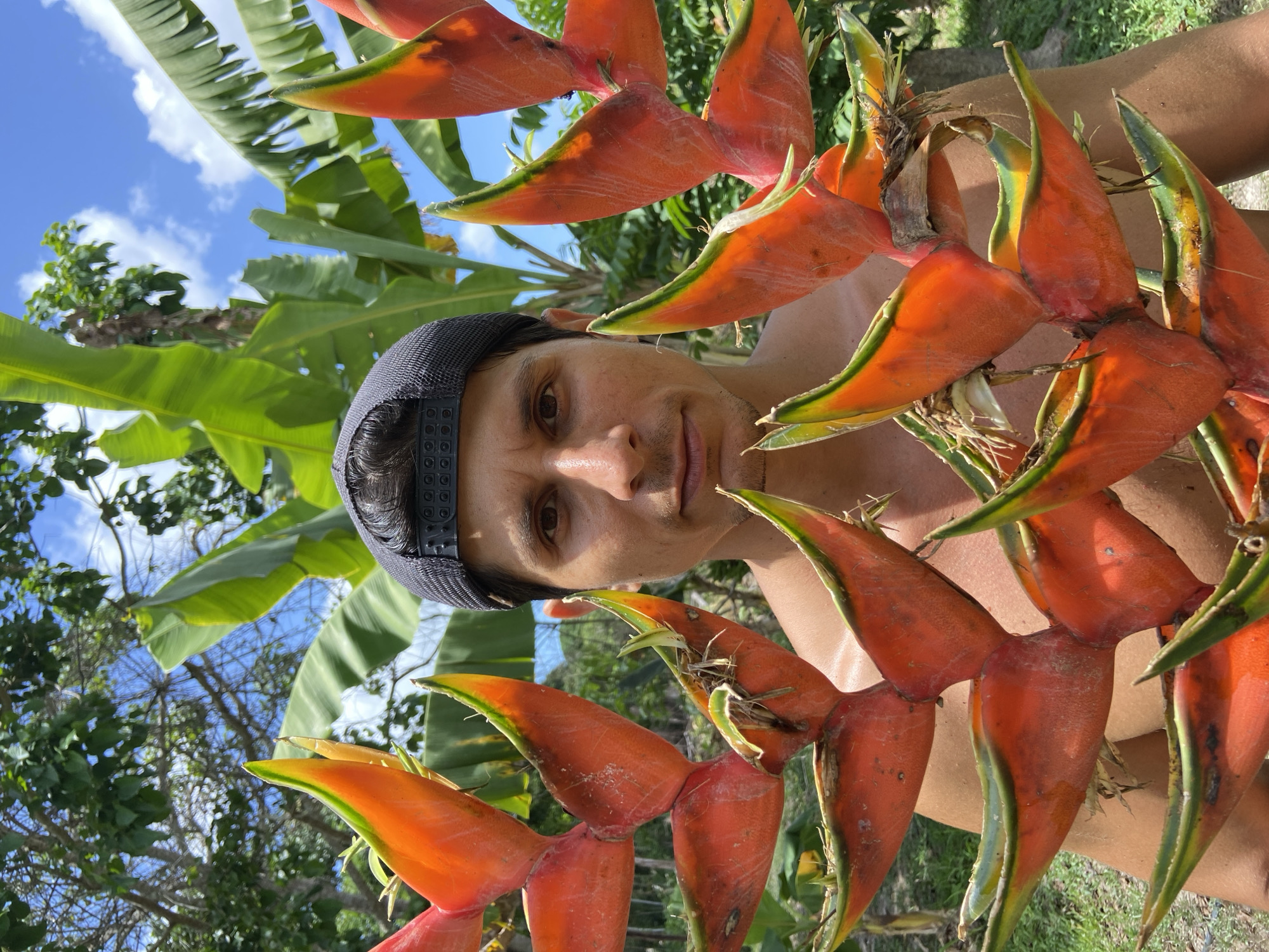 A medium shot photograph taken outdoors, likely in a tropical environment, features a man wearing a dark baseball cap and no shirt, positioned behind a large, vibrant Heliconia flower. The man's face is centrally framed, with his gaze directed towards the camera. The Heliconia, characterized by its bright orange and green bracts, is held in front of him, partially obscuring his body. Lush green foliage and banana leaves form the background, with a bright blue sky and white clouds visible above. The setting suggests a location in the city of Los Velásquez, Venezuela, characterized by its tropical climate and abundant vegetation.