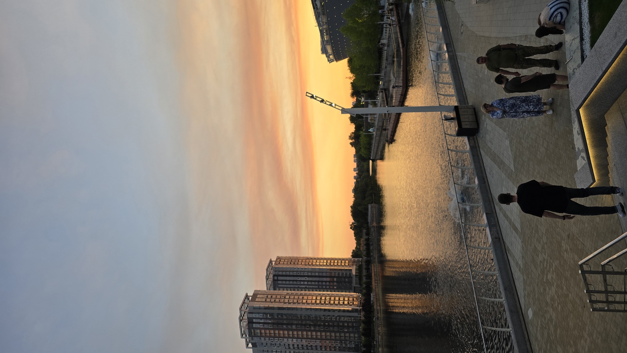 This image captures a serene evening scene in Kyiv, Ukraine, during sunset. The sky is painted with warm hues of orange and pink, reflecting beautifully on the calm water of a canal or river. Two modern high-rise buildings stand prominently in the background, their windows catching the golden light of the setting sun.
Along the paved walkway beside the water, several individuals are present. One person, wearing a black t-shirt and jeans, is walking away from the camera. Further along, a group of four people are visible. One person, wearing a blue and white patterned dress, is standing, while another, in military-style camouflage, stands nearby. A person in a dark t-shirt and shorts is also present, as is another individual bent over, partially obscured.
The atmosphere appears peaceful, with the soft glow of the sunset creating a tranquil mood. The time of day is clearly evening, as indicated by the sunset colors and the light conditions. The location cues, such as the architecture of the buildings and the general urban setting by the water, suggest a developed area within Kyiv. There is no visible text in the image.