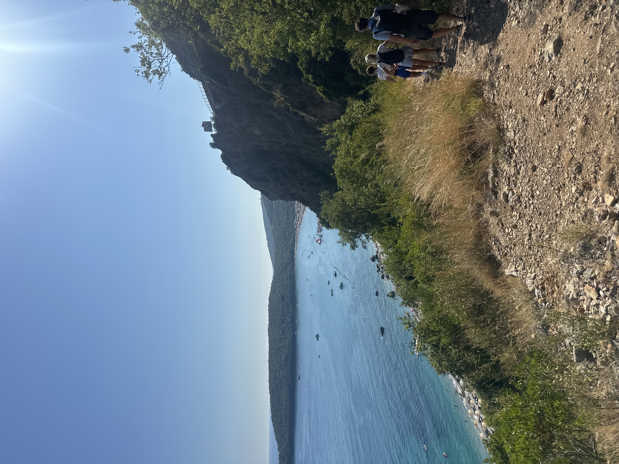 In Prijevor, Montenegro, a group of four individuals is walking along a rocky, unpaved path. The path is situated on a hillside overlooking a bay filled with turquoise water. Numerous boats are scattered across the water, and a beach lined with umbrellas and sunbathers is visible along the coastline. Dense green vegetation covers the slopes of the hills. The sky is clear and blue, with the sun prominently shining from the upper right corner, creating lens flare. The people are dressed in casual summer clothing and appear to be hikers, with two of them carrying backpacks. The overall scene depicts a scenic coastal landscape under bright sunlight.