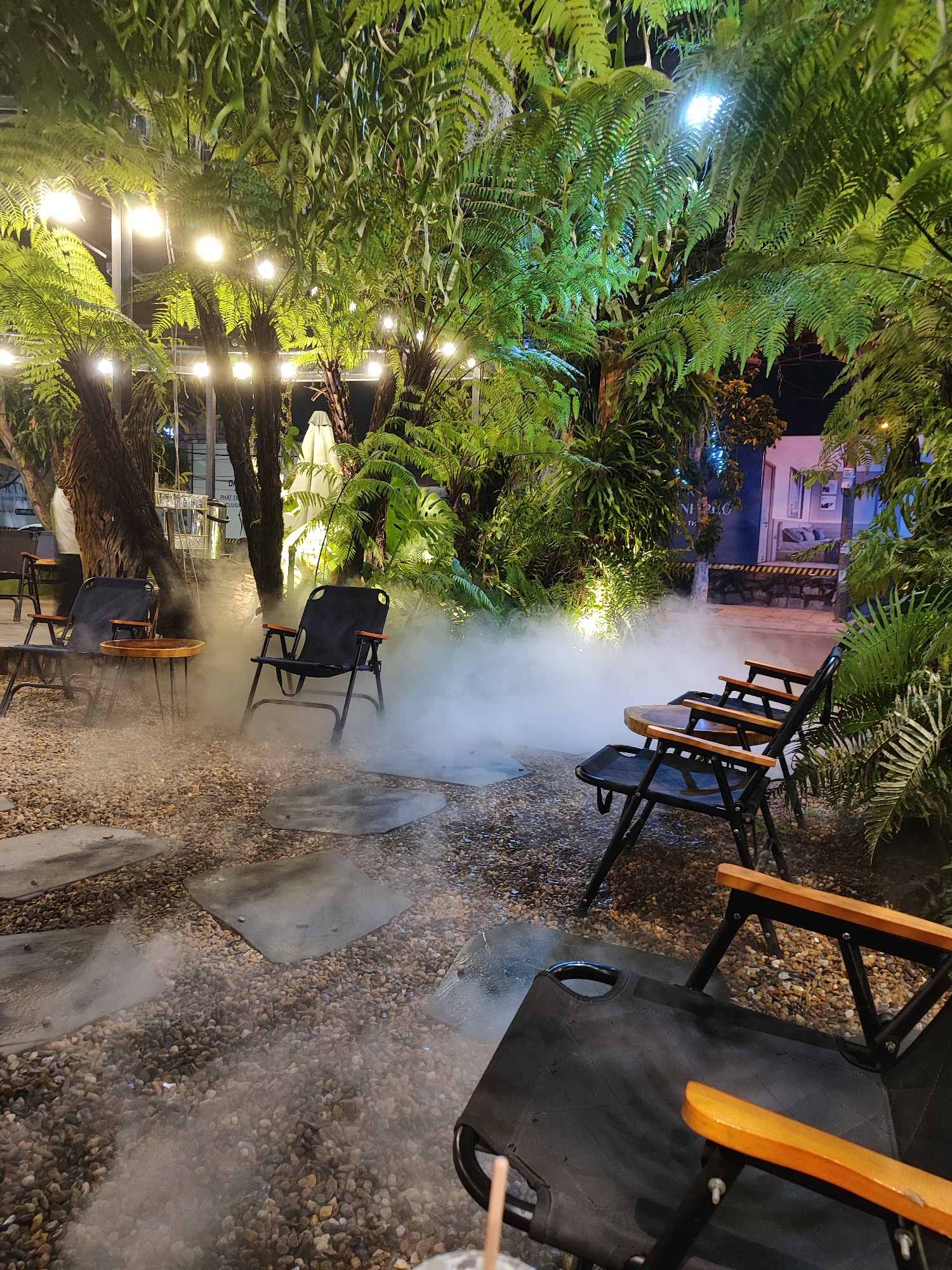 This image depicts an outdoor seating area, likely in Đà Nẵng, Vietnam, based on the tropical foliage and the lighting. There are several black folding chairs with wooden armrests arranged around small round tables. The ground is covered in small gravel and interspersed with flat, irregularly shaped stepping stones. A thick fog or artificial mist permeates the scene, obscuring the lower portions of the chairs and tables. Lush ferns and other tropical plants surround the seating area, with strings of lights hanging above. In the background, a building with a painted mural or image is visible. The scene appears to be set at night, illuminated by the overhead string lights and additional spotlights illuminating the foliage. No people are directly visible. The overall atmosphere is calm and inviting.