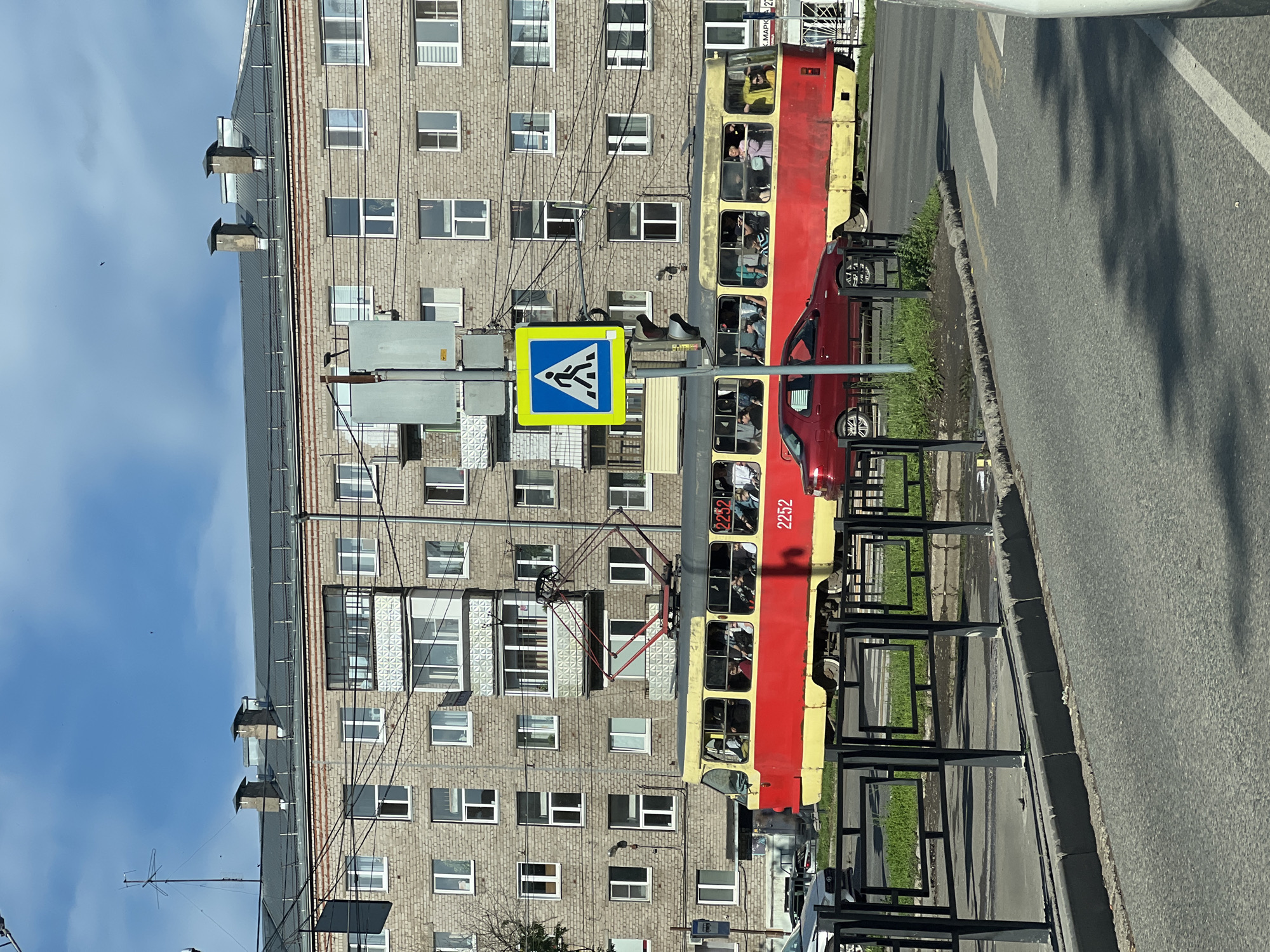 In Izhevsk, Russia, on a sunny day, a red and yellow tram with the number 2252 is traveling down a street. The tram, filled with passengers visible through its windows, is passing by a pedestrian crossing sign. To the left of the tram, a red car is parked. Behind the tram and car is a multi-story apartment building constructed of beige brick, with numerous windows and balconies. Power lines are visible running across the front of the building. The street in front of the tram and car is paved, with a sidewalk and black metal fencing. The sky is blue with some clouds.