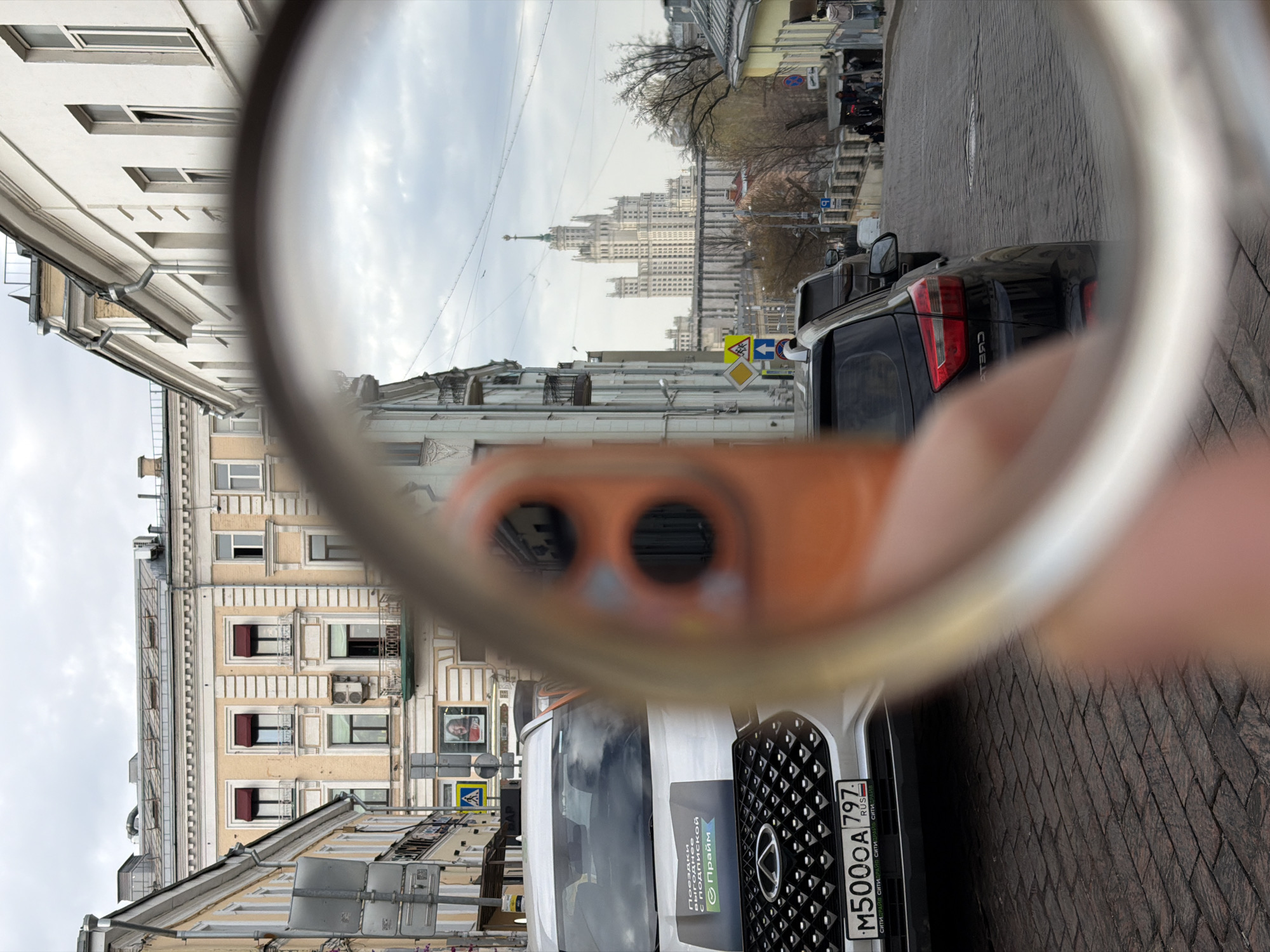The image is taken on a street in Moscow, Russia, where a person is holding a circular magnifying glass or lens to capture a view of the cityscape. The lens creates a distorted perspective, focusing on the buildings in the background while the surrounding street elements and the photographer's hand are blurred. A modern skyscraper is visible in the distance through the lens. In the foreground, a white car with a "Chery" emblem has a license plate that reads "M500OA 797 RUS." Street signs are evident, and buildings with architectural details and windows line the street. The weather appears overcast.