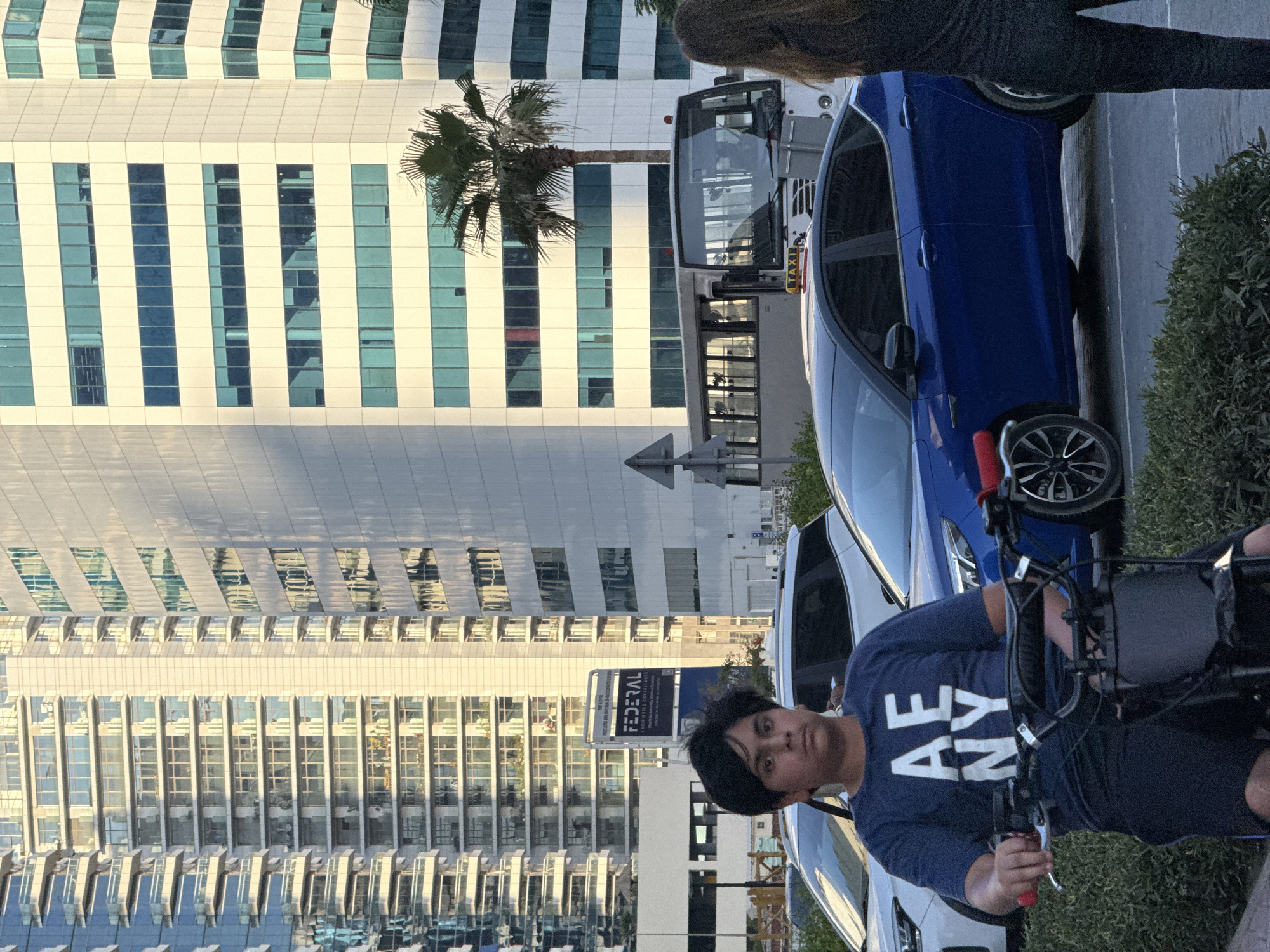 The image depicts a street scene in Dubai, United Arab Emirates. In the foreground, a young person is seated on a black electric scooter, wearing a dark blue t-shirt with "AE NY" printed in white. The person is facing forward, with a neutral expression. Behind the scooter, a blue sedan is parked. To the left of the blue sedan, a white SUV is visible. In the mid-ground, a light-colored bus is driving, with "TAXI" displayed on its roof. Further in the background, several modern skyscrapers with glass facades and balconies dominate the skyline. A palm tree stands to the right of the bus. The lighting suggests it is daytime, with sunlight casting shadows on the buildings. The scene appears to be in an urban area, likely a business district given the presence of tall office buildings.