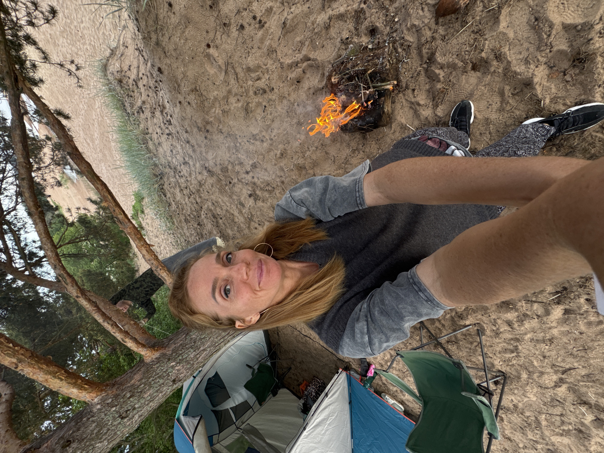An outdoor selfie captures a woman camping in a sandy, tree-lined environment in Zelenaya Roshcha, Russia. The woman, positioned centrally, has reddish-blonde hair, fair skin, and wears a grey two-tone long-sleeved top, patterned leggings, and black and white sneakers, along with gold hoop earrings. She holds the camera with an extended arm, smiling slightly. To her immediate left, two camping tents are pitched on the sand: a larger blue and white tent partially open to reveal a green folding chair, and a smaller white and blue tent. A large tree with thick, gnarled reddish-brown bark and pine needles dominates the left side, partially obscuring a second individual in a camouflage-patterned shirt, who appears to be handling a white rope or line near the tree trunk. To the woman's right, a small campfire burns on the sandy ground, producing bright orange flames and smoke from collected kindling and pine cones. The ground consists of light-brown sand mixed with scattered pine needles, dry grass, and debris. In the upper right background, a sloping expanse of light-colored sand leads down to a narrow, lighter strip, indicative of a beach bordering a body of water.
