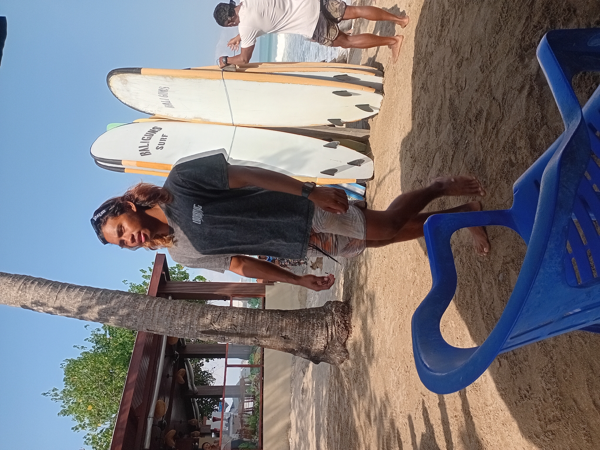 The image depicts a beach scene in Kuta, Indonesia, with a focus on a young man standing on the sand in front of several surfboards. The man, with long brown hair, wears a dark t-shirt and patterned shorts. He is looking to his left, with a slight smile. Behind him, two surfboards are leaning against a wooden structure, with text that appears to read "BALIGUNS SURF" on one of them. Another person, wearing a white t-shirt and patterned shorts, is visible to the right, near the water, interacting with one of the surfboards. The background shows a beach with sand, a palm tree, and a building with a thatched roof, likely a beach bar or restaurant. The ocean is visible in the distance, with waves gently rolling in. A blue plastic chair is in the foreground, partially obscuring the sand. The scene is set on a sunny day, indicated by the bright lighting and clear blue sky.