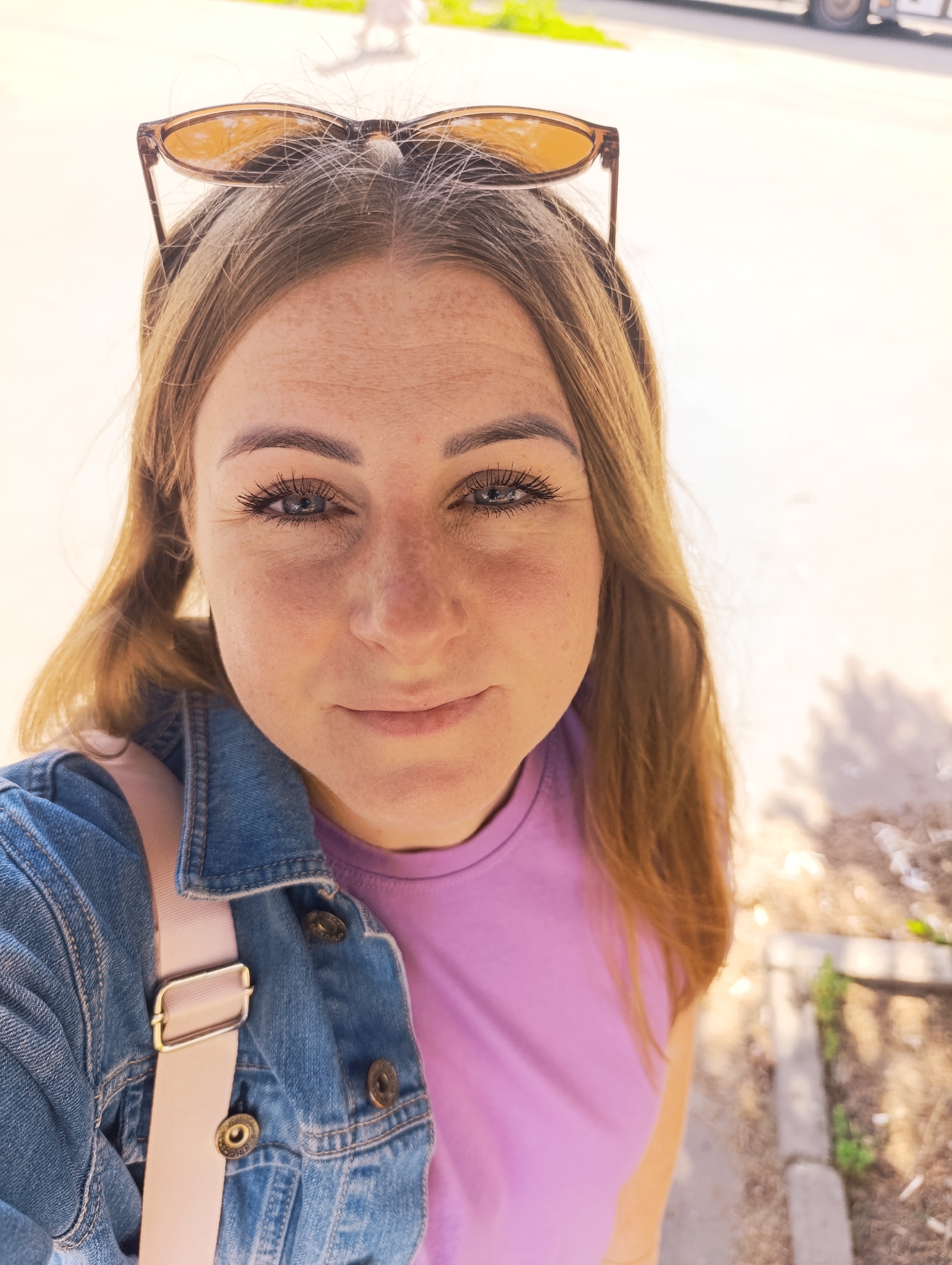 A close-up selfie of a young woman with long, blonde hair. She is wearing a denim jacket over a lavender t-shirt, and sunglasses are perched on top of her head. The woman has blue eyes and light skin with freckles. She is smiling subtly and looking at the camera. A light pink strap from a bag is visible across her chest.
The background is out of focus, but appears to be an outdoor setting with sunlight casting shadows. There is a curb and some dry leaves or debris on the ground, suggesting it might be a sidewalk or park area. The lighting indicates it is daytime, likely sunny. The overall mood of the image is casual and relaxed. There is no visible text in the image.