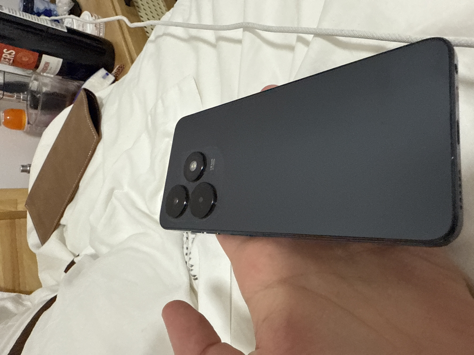 An overhead shot, taken in Haizhu District, China, shows a person's hand holding a dark grey smartphone on a white, creased bed. The phone's rear camera module, featuring three circular lenses and a flash, is prominently displayed. The background includes a wooden headboard, a brown leather case, and various items on a bedside table, such as a bottle of wine and glass perfume bottles. A white charging cable snakes across the bed.