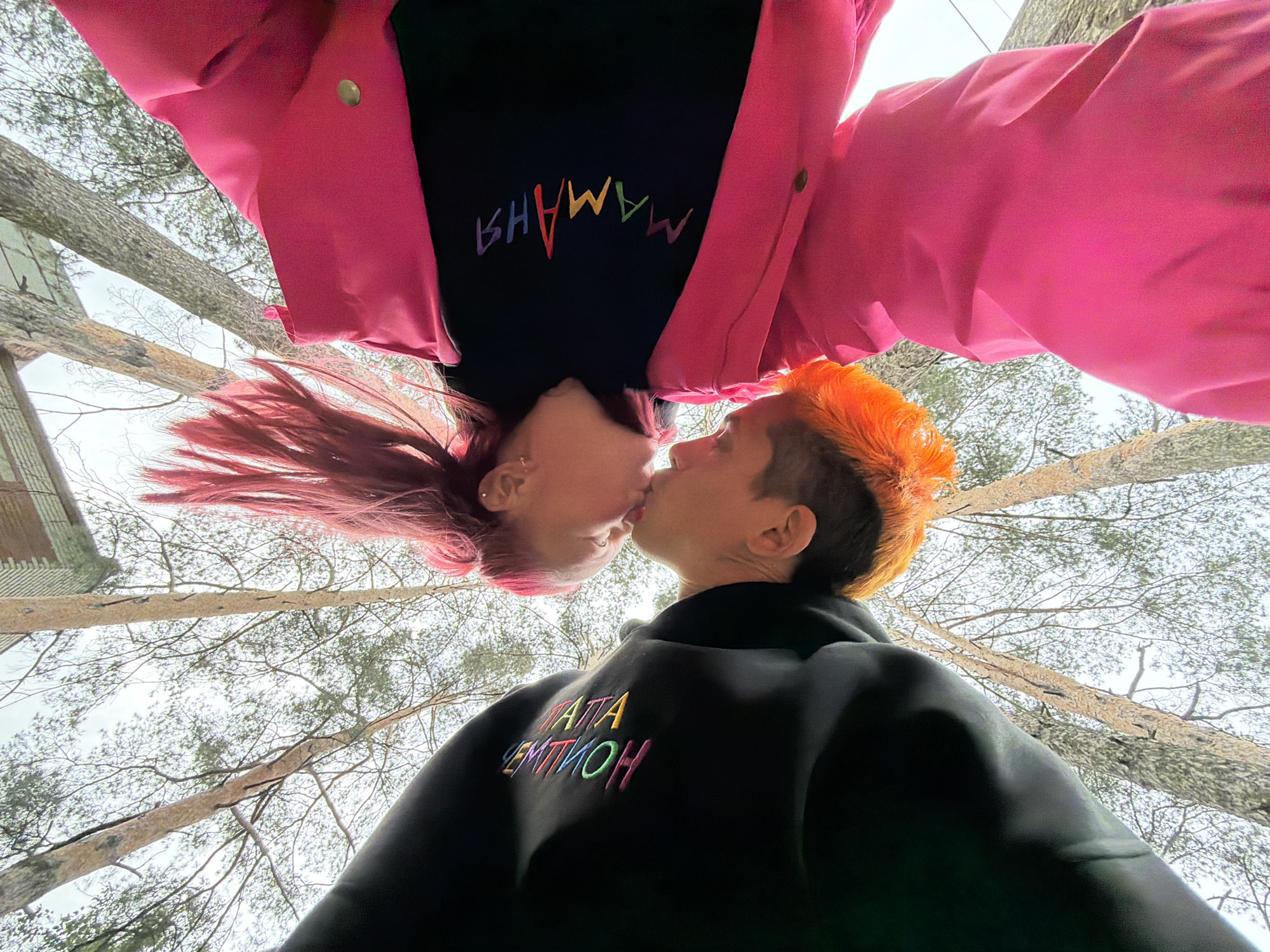 This low-angle, upward-looking photograph captures two individuals in a kissing embrace, positioned at the bottom of the frame against a backdrop of tall trees and an overcast sky in Chaikovsky, Russia. The individual on the left has short, bright orange hair and wears a black long-sleeved garment featuring "ПАПА АНТОН" embroidered in rainbow-colored Cyrillic letters across the chest. The individual on the right has long, bright pink hair, styled upwards, and wears a black top under a vibrant pink jacket, with "МАМАНЯ" embroidered in rainbow-colored Cyrillic letters across their chest. Their faces are close, lips touching, with their bodies largely obscured by the camera's angle. In the upper half of the image, numerous bare or sparsely foliated trees with light-colored trunks extend vertically into a bright, white sky. A portion of a building with a textured, possibly brick or stone, facade and a sloped roof is visible in the upper right background, situated behind the trees.