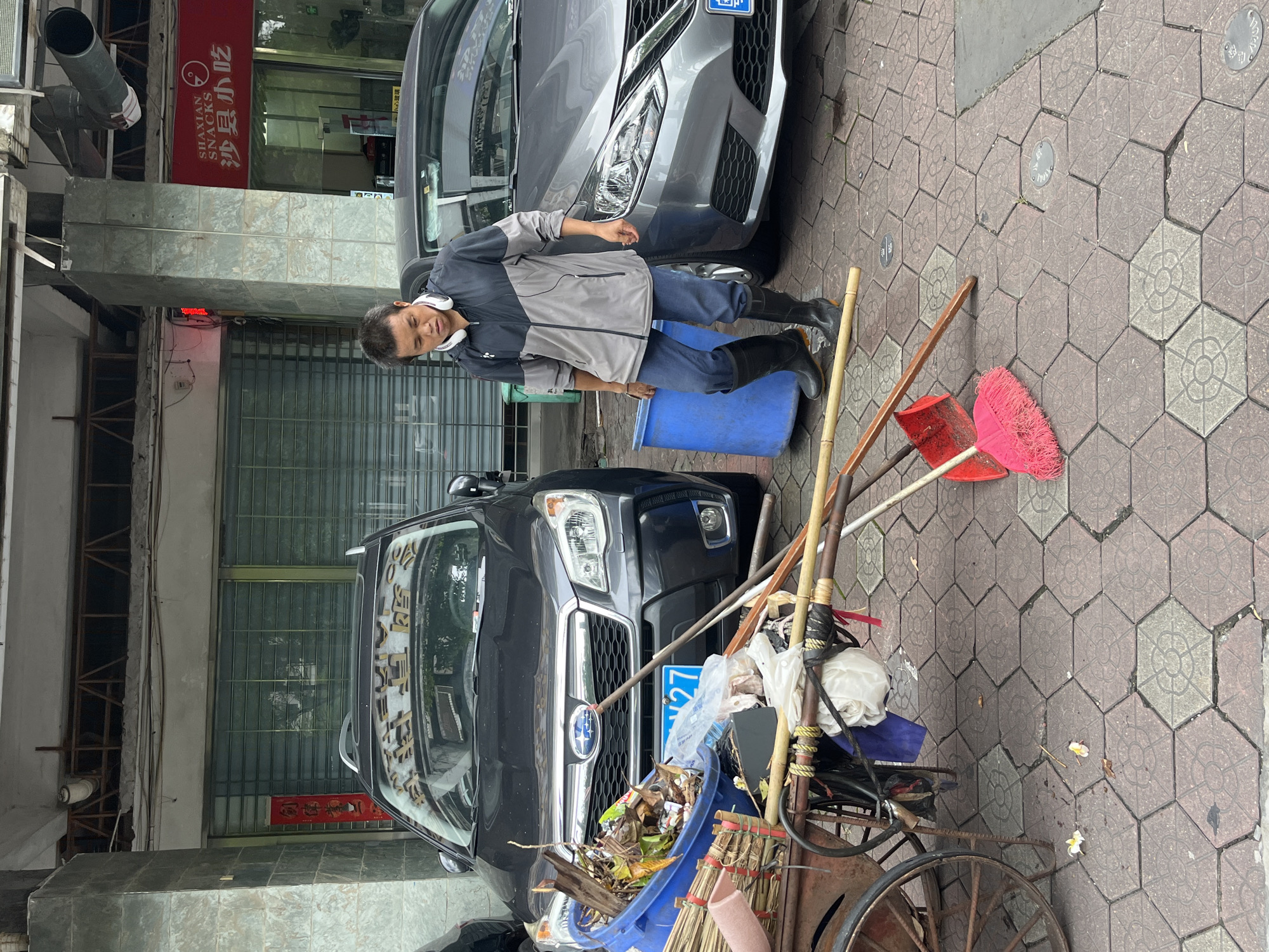 A man wearing a grey and black jacket, blue pants, and black boots stands on a hexagonal-tiled sidewalk in front of a building with a grey tiled facade and a closed metal grate. Next to him is a blue plastic bin and a cart filled with debris and cleaning tools. Two cars are parked behind the cart, a dark grey Subaru Forester on the left and a silver sedan on the right. The Subaru has a blue license plate with the number "X27" visible. The man is looking down at the cart. In the background, a red sign with white Chinese characters reads "SHAXIAN SNACKS" and "沙县小吃". The scene appears to be a street in the city of Chancheng District, China.