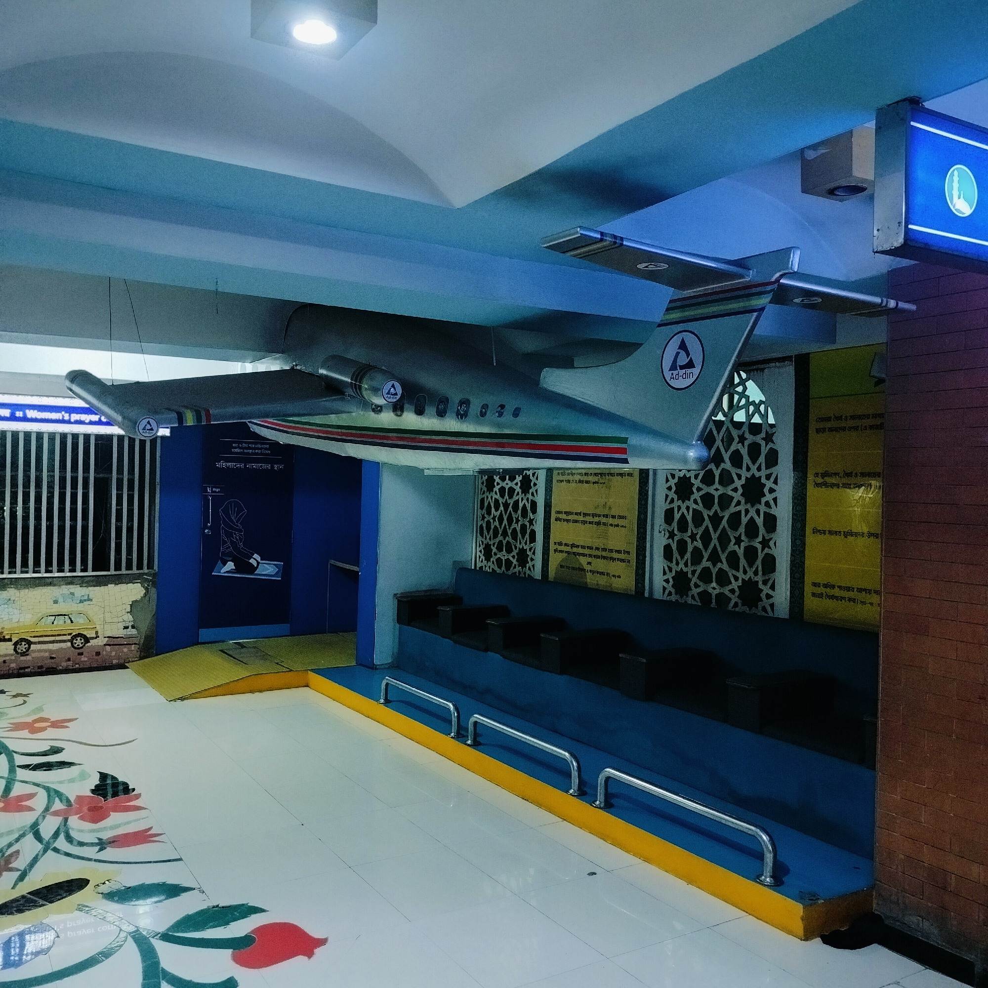 The image shows an interior space, likely in Dhaka, Bangladesh, featuring a model of an airplane suspended from the ceiling. The aircraft has the words "Ad-din" on the tail and engine. Below the plane, there is an elevated platform with seats, possibly for prayer or resting. There are signs in Bengali, including one indicating "Women's prayer." Decorative elements include geometric patterns on a wall and a floral design on the floor. A small mural with a depiction of a car is visible in the background. The lighting is artificial and the environment appears to be indoors.