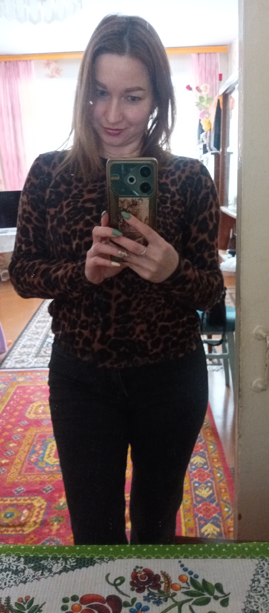 A person stands in front of a mirror and takes a selfie. The person has long brown hair and is wearing a brown and black leopard print sweater and black jeans. The phone being used to take the photo is a dark color with multiple lenses on the back. The person has manicured nails that are painted green. The scene is indoors in a room that is somewhat cluttered. In the background there is a rug with a red and yellow pattern. The city and exact location is unknown.