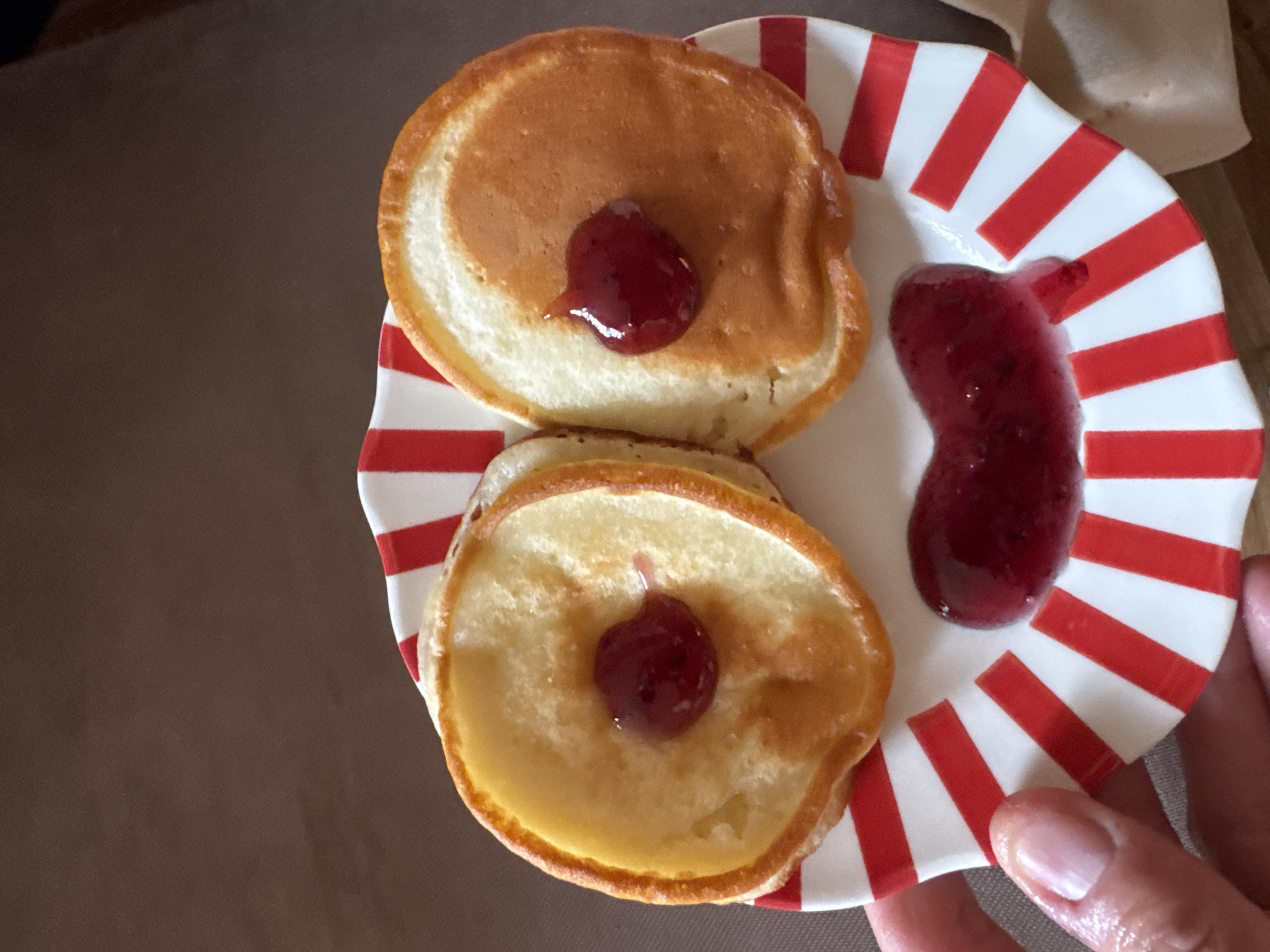 A person is holding a plate of pancakes decorated to resemble a face. Two pancakes serve as eyes, each with a dollop of jam in the center. A smear of jam forms a smile below the eyes. The plate has a red and white striped pattern. The background is a blurred brown surface.