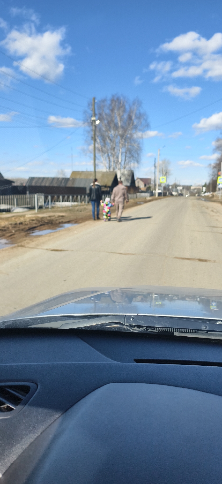 The image shows a view from inside a vehicle, looking out onto a road in Фоки, Russia. In the distance, two adults and a child are walking along the right side of the road. The adult on the left is wearing a dark jacket and jeans, while the adult on the right is wearing a tan jacket. The child is wearing a colorful jumpsuit. Buildings are visible on either side of the road. The sky is blue with white clouds.