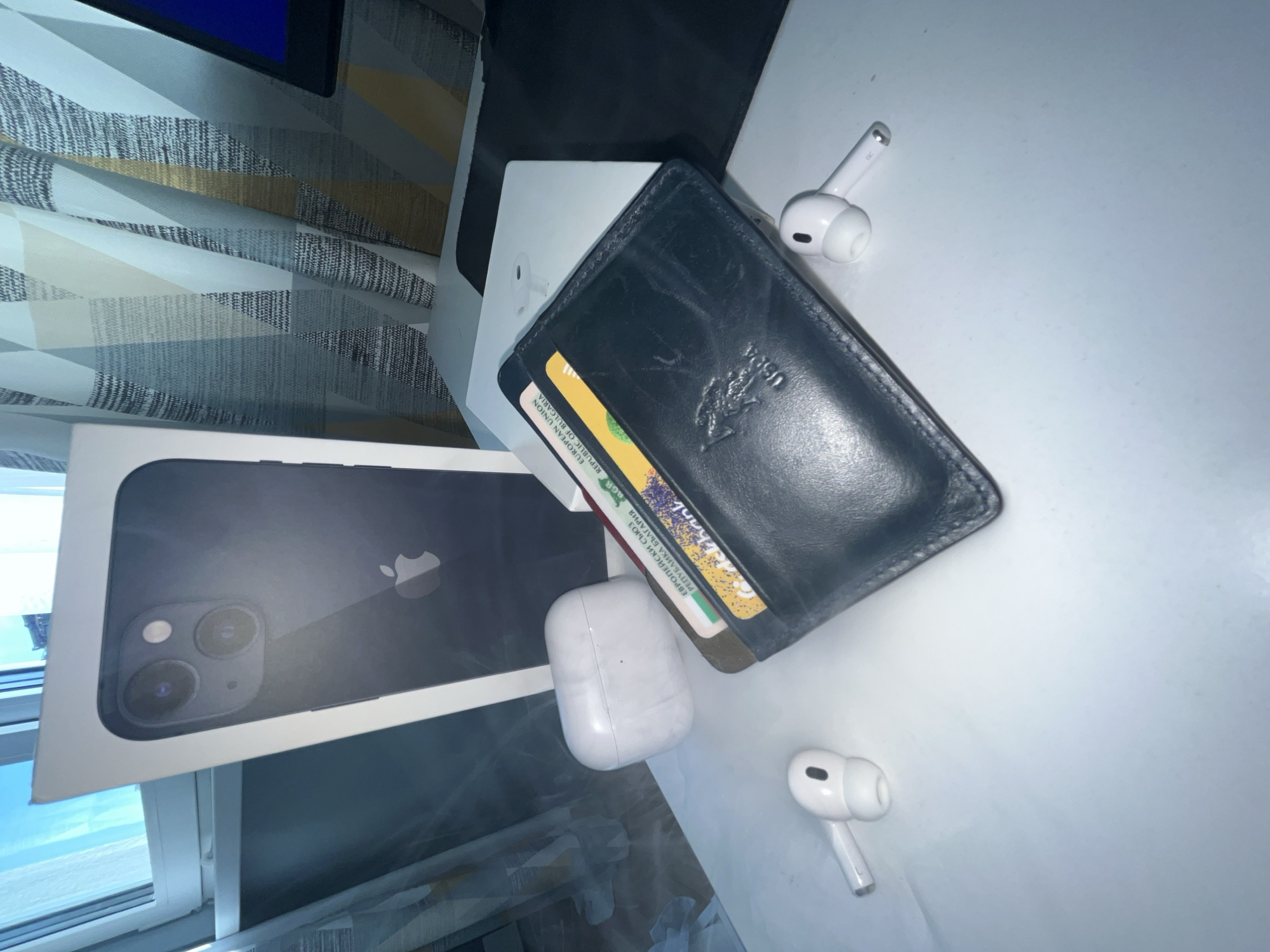 On a white surface, an iPhone box, a black leather wallet, and a pair of white AirPods are arranged. A curtain with a geometric pattern is visible in the background. The location appears to be indoors, possibly in Veliko Tarnovo, Bulgaria based on the currency card in the wallet.