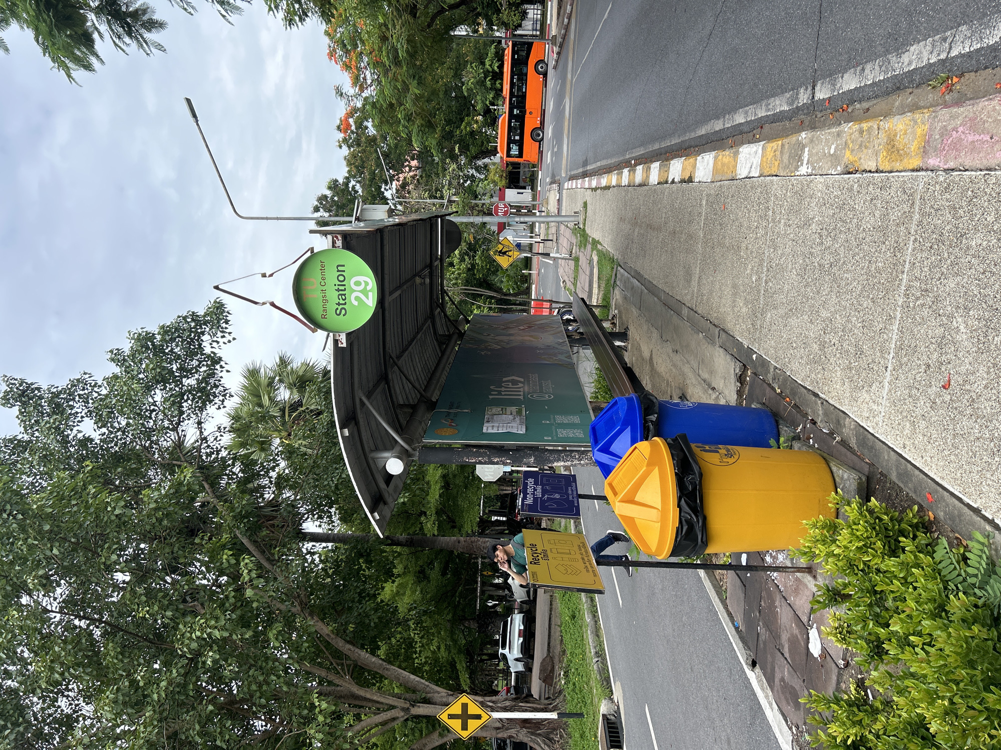 The image depicts a bus stop in Tha Khlong Town Municipality, Thailand. A green circular sign with "Rangsit Center Station 29" is visible above the bus stop shelter. The shelter has a metal roof and a bench. In front of the shelter are two trash bins, one yellow and one blue, and two recycling signs. A person in a green shirt is standing near the recycling signs. A paved sidewalk runs alongside the road, with a strip of grass and trees on the other side. An orange bus is driving on the road. The sky is overcast.