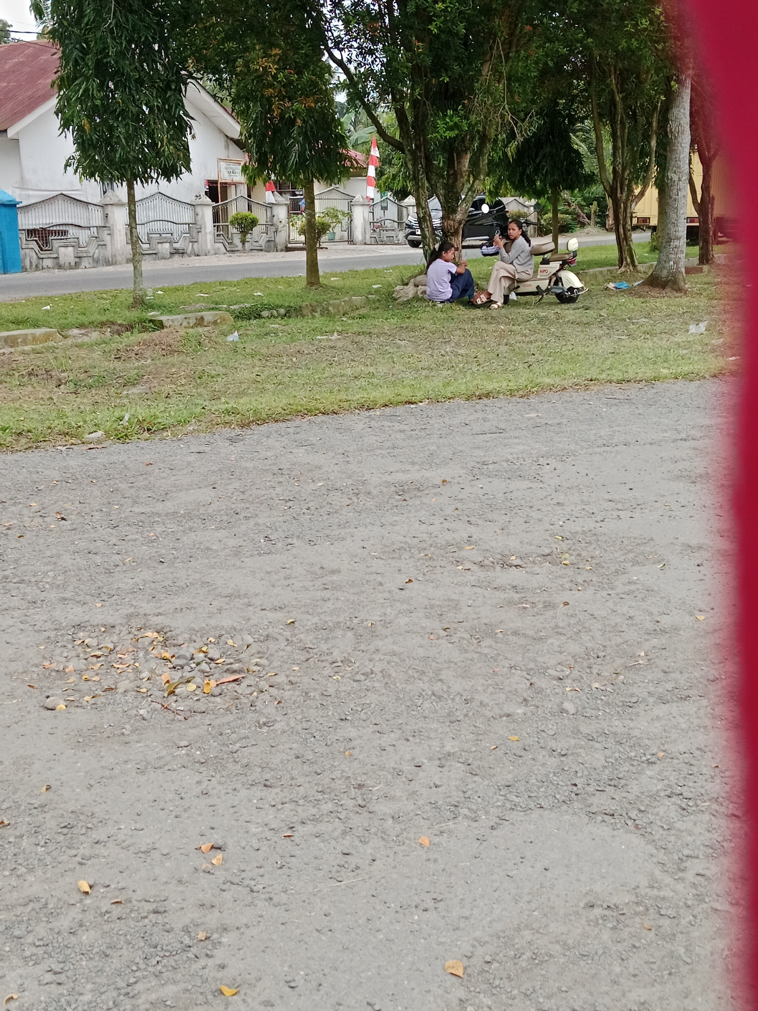 Two young women are seated on a grassy area next to a paved road. One woman, wearing a light-colored long-sleeved shirt and pants, is sitting on a beige scooter or motorcycle. The other woman, wearing a light purple shirt and dark pants, is sitting on the grass next to her. They appear to be in conversation. A white helmet is visible on the back of the scooter. The background shows a building with a red roof and white walls, a fence, several trees, and a street with a few parked vehicles. The setting appears to be a public area, possibly a park or a roadside rest stop, in a tropical climate given the lush greenery. The time of day seems to be late afternoon, with soft lighting. The overall mood appears relaxed and casual. The image is a photograph taken from a slightly low angle. A bright red vertical stripe is visible on the right edge of the frame, suggesting it may be part of a larger banner or a frame element. No legible text is visible in the image.