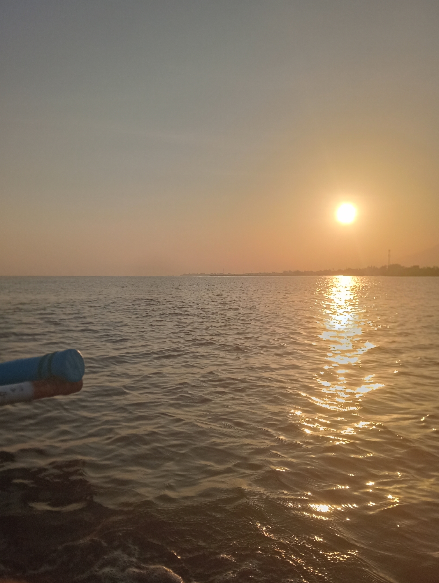 A serene sunset over the calm waters of the sea in Besuki, Indonesia. The sun, a bright orb, is positioned in the upper right quadrant of the image, casting a shimmering reflection across the water's surface. The sky transitions from a pale blue at the top to a warm, hazy orange near the horizon. The silhouette of the distant coastline is visible, dotted with faint structures and what appears to be a communication tower. In the foreground, the edge of a boat is visible on the left, with a blue and white object protruding into the frame. The water exhibits gentle ripples, reflecting the warm glow of the setting sun. The overall atmosphere is tranquil and peaceful.
