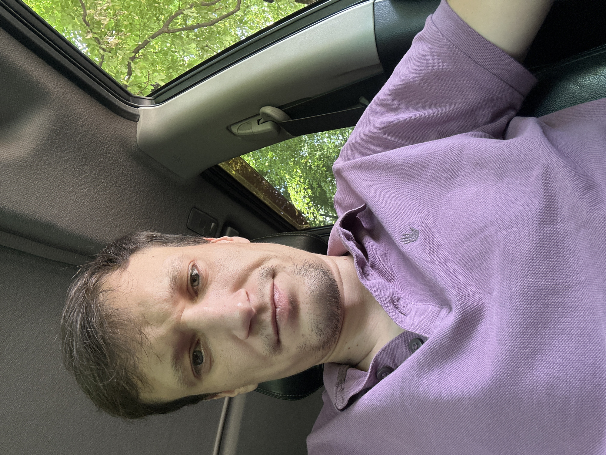 A close-up, outdoor, eye-level, selfie shot shows a fair-skinned man in his late 20s or early 30s sitting in the driver's seat of a vehicle. He has medium-brown, thinning hair, light-brown eyes, and light stubble on his face. He is wearing a purple polo shirt with a small grey embroidered logo on the left chest. The man is looking directly at the camera with a slightly concerned or questioning expression. Behind him, out the car window, is lush green foliage and trees, indicating an outdoor setting with abundant vegetation. The interior of the car is visible with a grey headliner, part of the driver's side pillar, and a black seat. The location of Shatava, Ukraine is not discernible from the image.