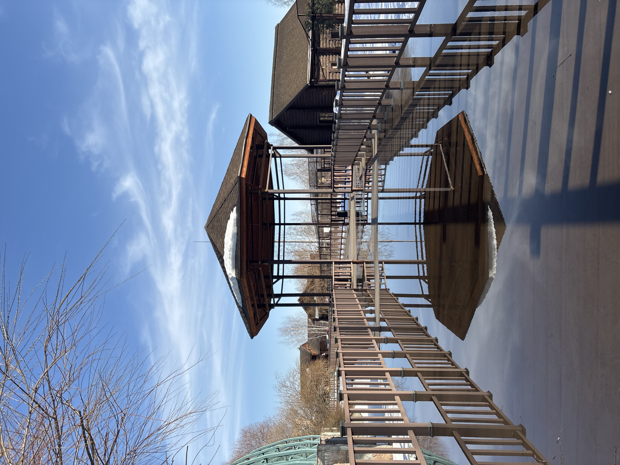 The image shows a building reflected in a body of water under a blue sky with sparse clouds. The building has a wooden structure, including a peaked roof with some remaining snow. A covered walkway with railings is visible, also reflected in the water. Bare trees are in the foreground. The location is likely Izhevsk, Russia, based on visual cues of architecture and environment.
