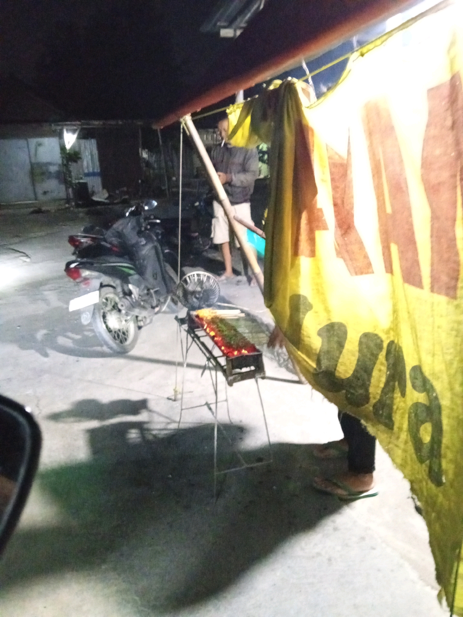 The image depicts an outdoor scene at night in Kedungwinong, Indonesia. A man, dressed in a grey jacket and white shorts, stands near a yellow and brown banner. In the foreground, a motorcycle is parked on a concrete surface. Adjacent to the motorcycle, a metal grill with food items is set up. The environment appears to be a street or an open area, possibly a food stall, with a makeshift roof structure extending overhead. The lighting suggests artificial illumination.
