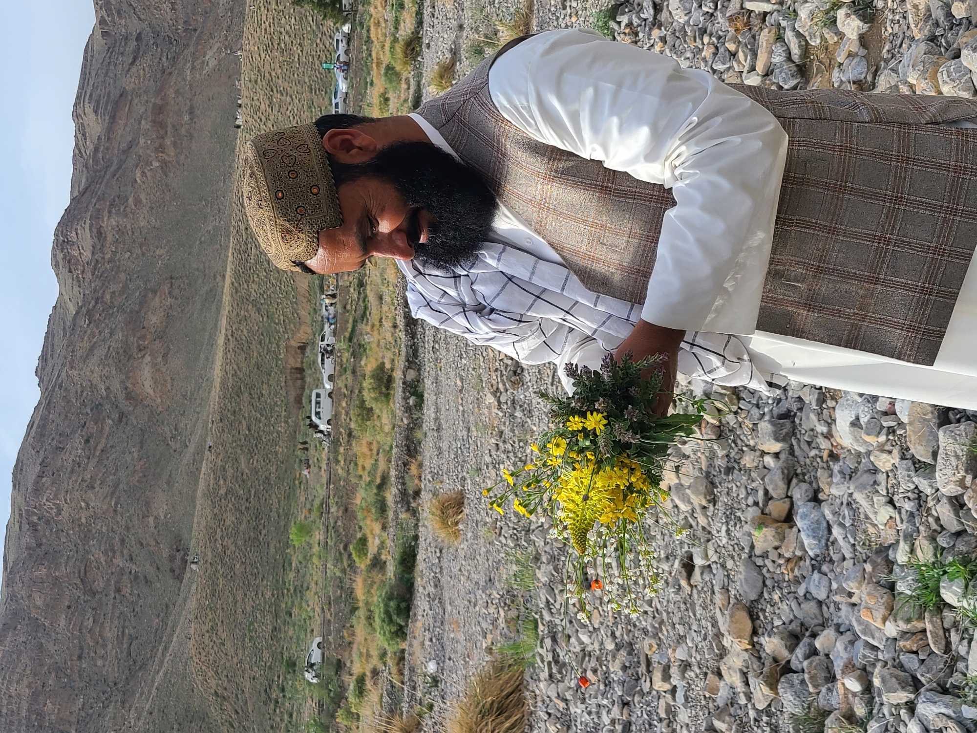 A bearded man stands on rocky terrain in Quetta City Tehsil, Pakistan, holding a bouquet of wildflowers. He is depicted in a three-quarter profile, looking down at the flowers with a slight smile. He wears a patterned, embroidered cap, a white long-sleeved garment, and a brown and white plaid vest. A white and black checkered cloth is draped over his left shoulder. The bouquet, held in both hands near his chest, consists primarily of bright yellow and white flowers, with some purple-hued sprigs and green foliage; two small orange flowers are also visible on the left side.
The immediate ground consists of numerous grey and brown rocks and pebbles, interspersed with sparse patches of dry grass and small green plants. In the mid-ground, a dirt road or track is visible with several parked white vehicles, including sedans and vans, and multiple small groups of people. The background features extensive, arid, treeless mountains with sparse, brownish-green vegetation under a clear, light blue sky.