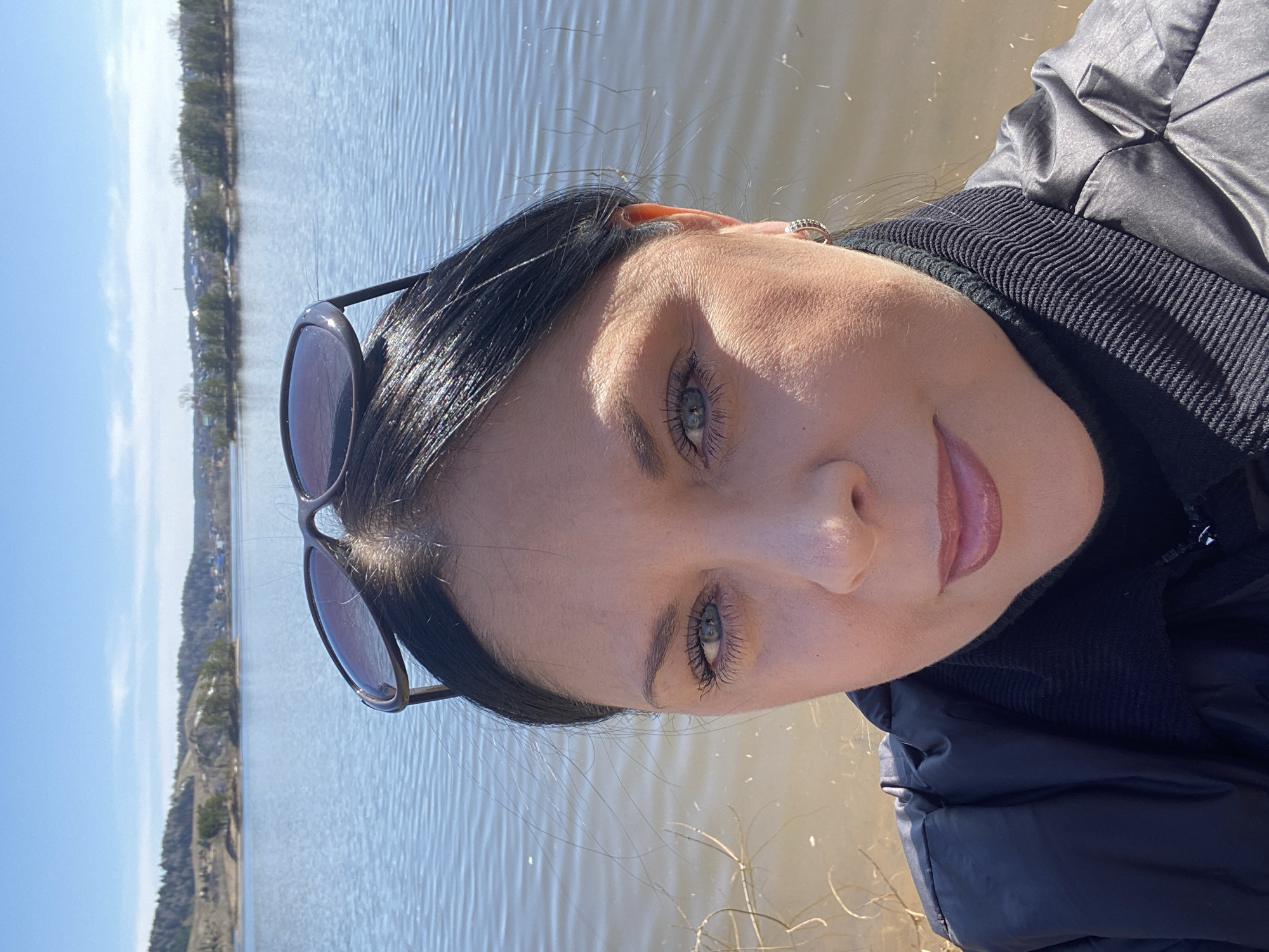 A woman is photographed outdoors on a sunny day, likely near a body of water. She has dark hair, fair skin, and light-colored eyes. Sunglasses are perched on her head. She is wearing a dark jacket and a black scarf or turtleneck. The background features a body of water and a distant shoreline with trees and buildings. The location could be the city of Noviy, Russia, based on a hypothesis.