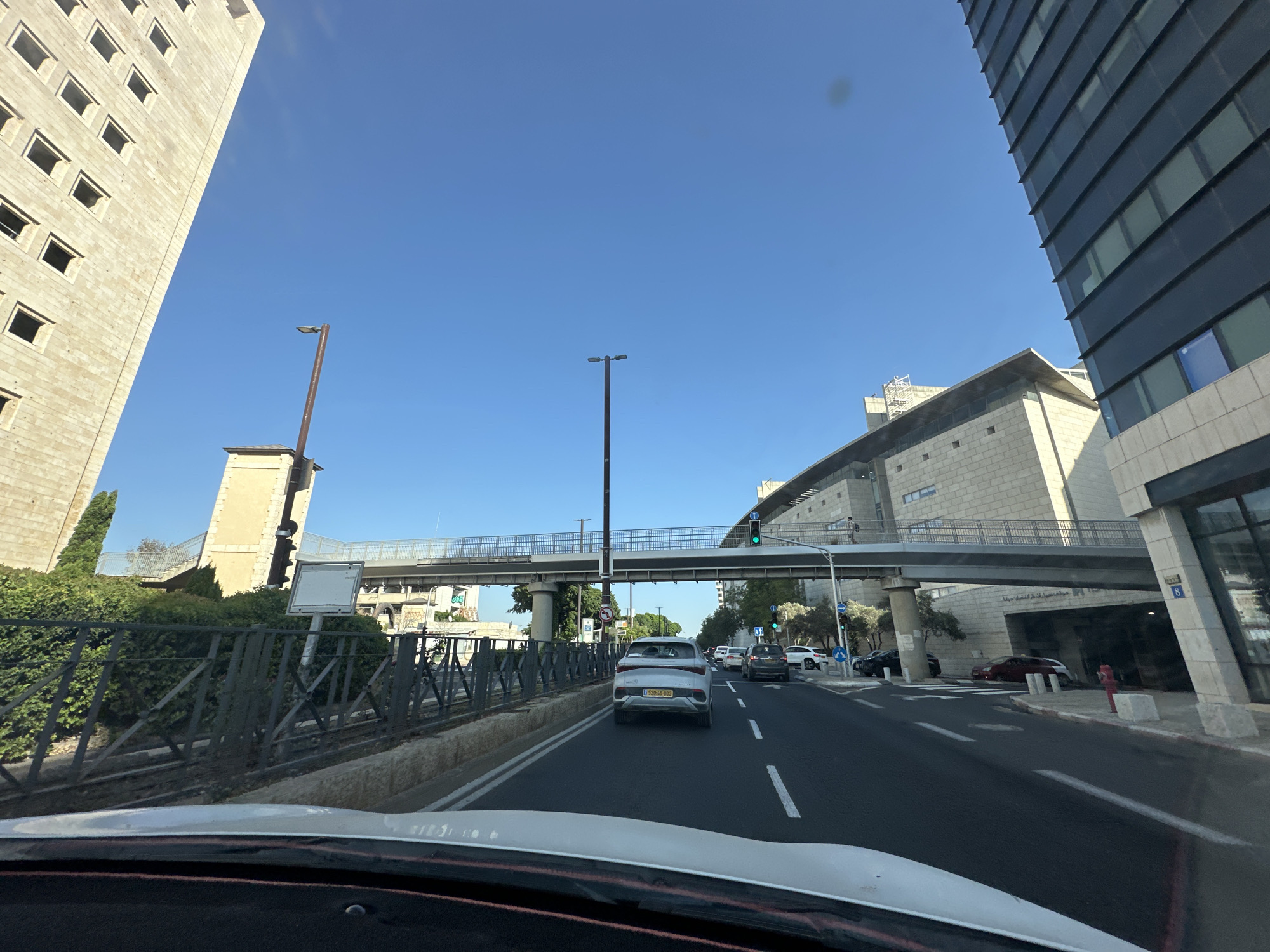The image captures a street view in Haifa, Israel, likely from a vehicle's perspective. The scene is dominated by modern architecture, with a tall, light-colored building with numerous rectangular window openings on the left, and a sleek, dark-glass building on the right. A pedestrian overpass with a metal railing spans across the road in the middle ground, connecting the buildings. The road is asphalt with white lane markings, and several cars are visible, including a white SUV in the foreground and a dark car behind it. Streetlights are present, and traffic lights are visible, with a green light illuminated above the pedestrian overpass. Lush green bushes line the side of the road behind a metal fence. The sky is a clear, bright blue, indicating daytime and fair weather. The overall impression is a contemporary urban environment with significant infrastructure.