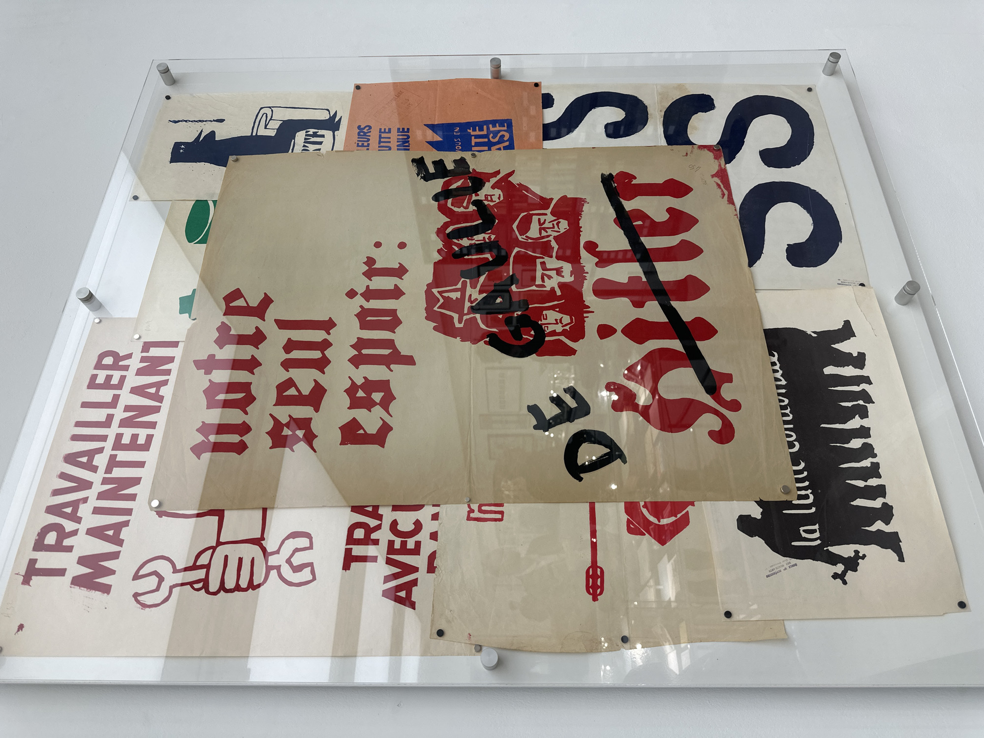 The image shows an arrangement of several posters, possibly protest or propaganda posters, layered and displayed within a transparent, mounted frame. The frame is attached to a white wall with visible metal standoffs.
Several posters are visible:
* A poster on the left with the words "TRAVAILLER MAINTENANT" (Work Now) and an image of a hand holding a wrench, printed in red ink.
* A poster with gothic-style text "notre seul espoir" (Our Only Hope) in red.
* A poster with red figures and the word "de" with a black mark through it and the word "Sitter" on it.
* Several partial visible posters showing graphic icons, such as "la lutte continu", "leurs utte inue" and blocky navy blue S shapes.
The posters are old and show signs of age, with creases and yellowing of the paper. The posters are arranged in a visually layered and slightly chaotic manner, giving a sense of historical context.
There is no indication of location in the city of London, United Kingdom.