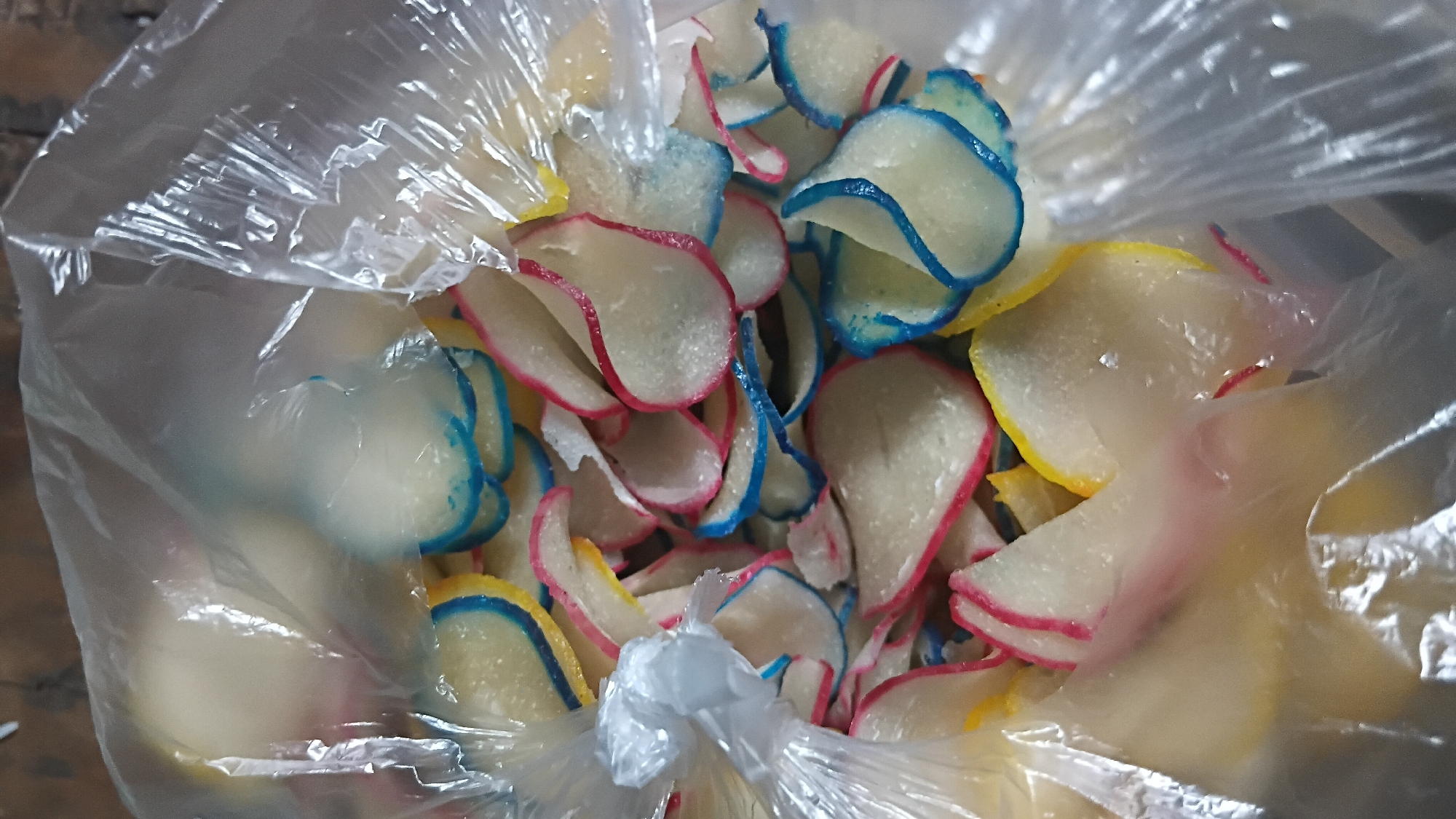 A clear plastic bag is filled with a colorful assortment of thin, wavy crackers. The crackers are mostly white with rims of vibrant colors, including red, blue, and yellow. Some crackers show a gradient of these colors. The bag is partially open, revealing the contents within. The background is dark and out of focus, suggesting the crackers are the primary subject. The image appears to be a close-up shot, likely taken indoors or in a controlled setting, as indicated by the lighting and the plastic bag. There are no people or discernible environmental elements that would place this image specifically in Prabumulih, Indonesia; it depicts a generic food item.