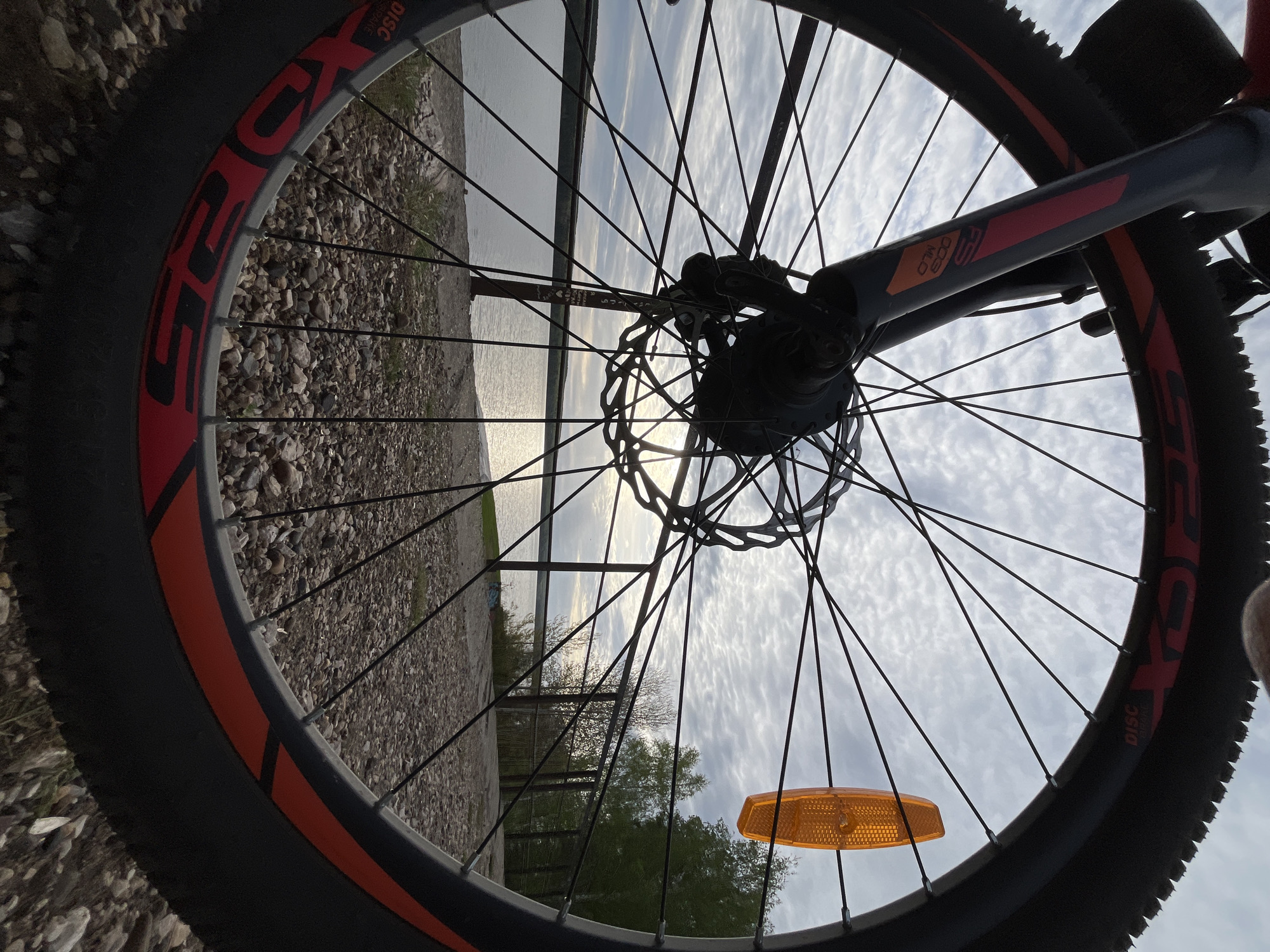 The image shows the front wheel of a bicycle, with the spokes radiating from the central hub. The disc brake is visible behind the spokes. An orange reflector is mounted on one of the spokes. The tire is black with a red accent stripe on the sidewall. The foreground is a gravelly or rocky surface. Through the wheel, the background shows a body of water with a shoreline visible on the left. A low fence runs along the shoreline, and trees are visible beyond it. The sky is cloudy. The location is in the city of Chaikovsky, Russia.