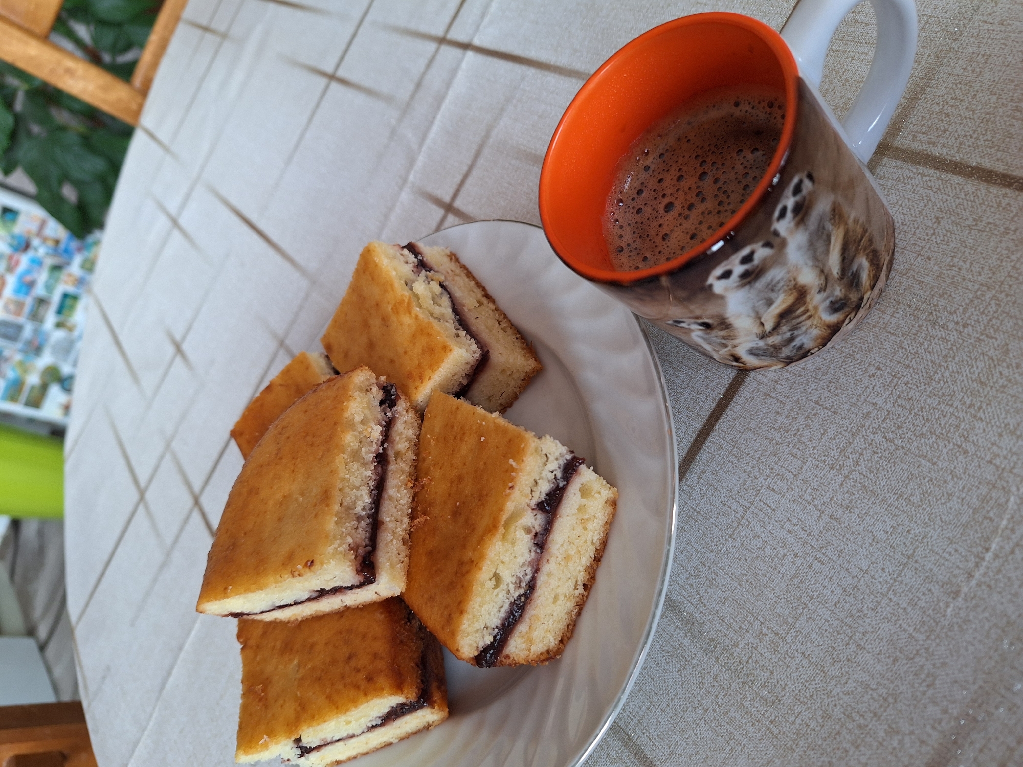 A plate of layered sponge cake with jam is arranged next to a mug of coffee. The mug is white with an orange interior and a print of meerkats. The plate and mug sit on a tablecloth with a grid pattern. In the background, a plant and a blue and white object are partially visible. The location cannot be determined.