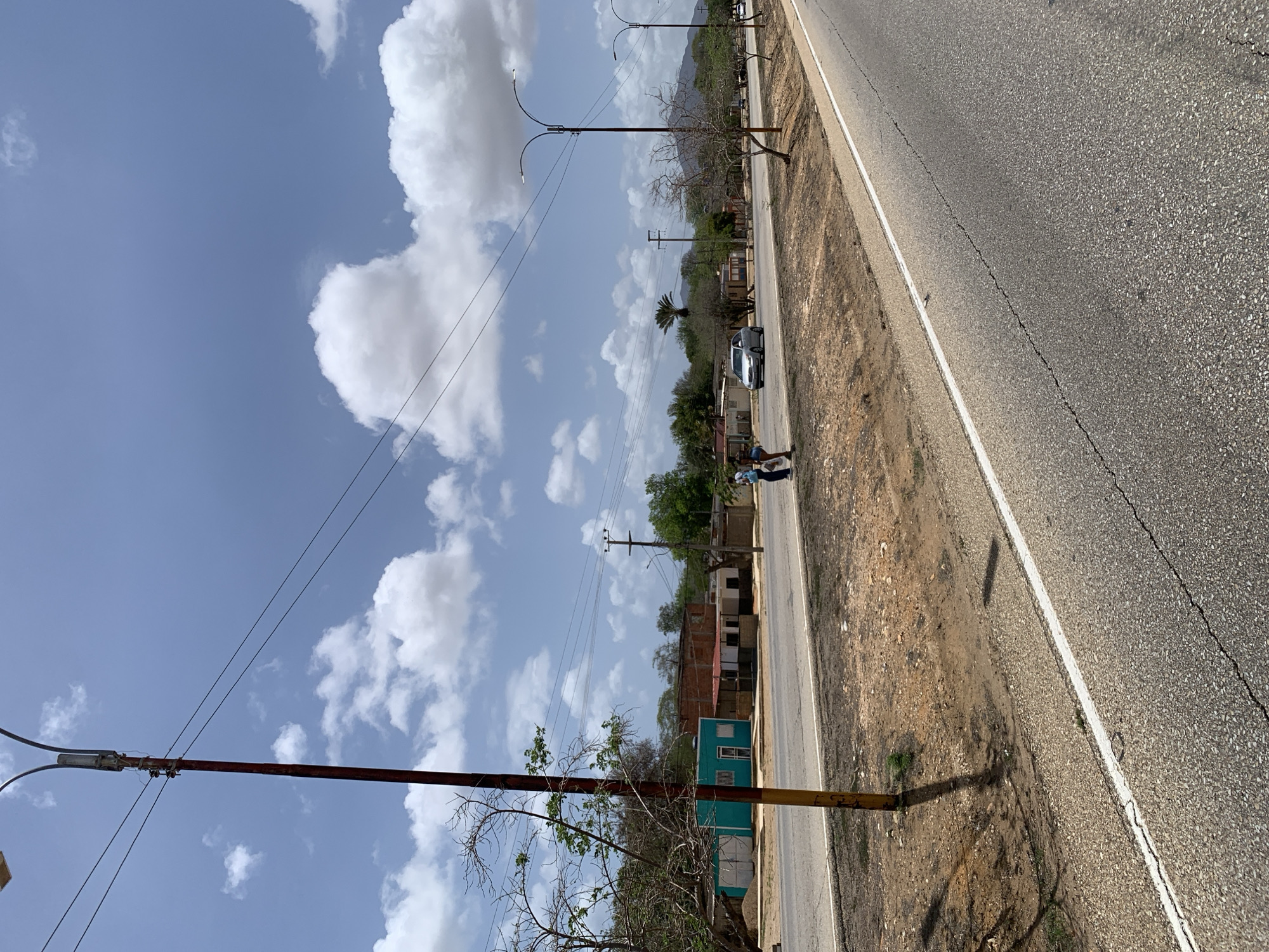 The image captures a scene along a paved road in El Dátil, Venezuela. The road runs diagonally from the lower right to the upper left of the frame. The surrounding environment is dry and arid, with patches of brown soil and sparse vegetation. Several buildings, appearing to be houses or small commercial establishments, are visible on the left side of the road. They are of various colors and architectural styles, with some appearing to be under construction or in disrepair.
Two individuals, a male and a female, are standing on the side of the road, near a silver sedan. The man appears to be wearing a white shirt and dark pants, while the woman is in a light-colored top and shorts. They are facing each other, and it is unclear if they are interacting. The car is parked on the side of the road, facing away from the viewer.
In the foreground, a tall utility pole with a light fixture stands prominently on the left side of the road. Power lines are strung across the image, connecting to other utility poles. The sky is a bright blue with scattered white clouds. The overall impression is that of a rural or semi-rural setting within El Dátil, Venezuela, under a clear, sunny sky.