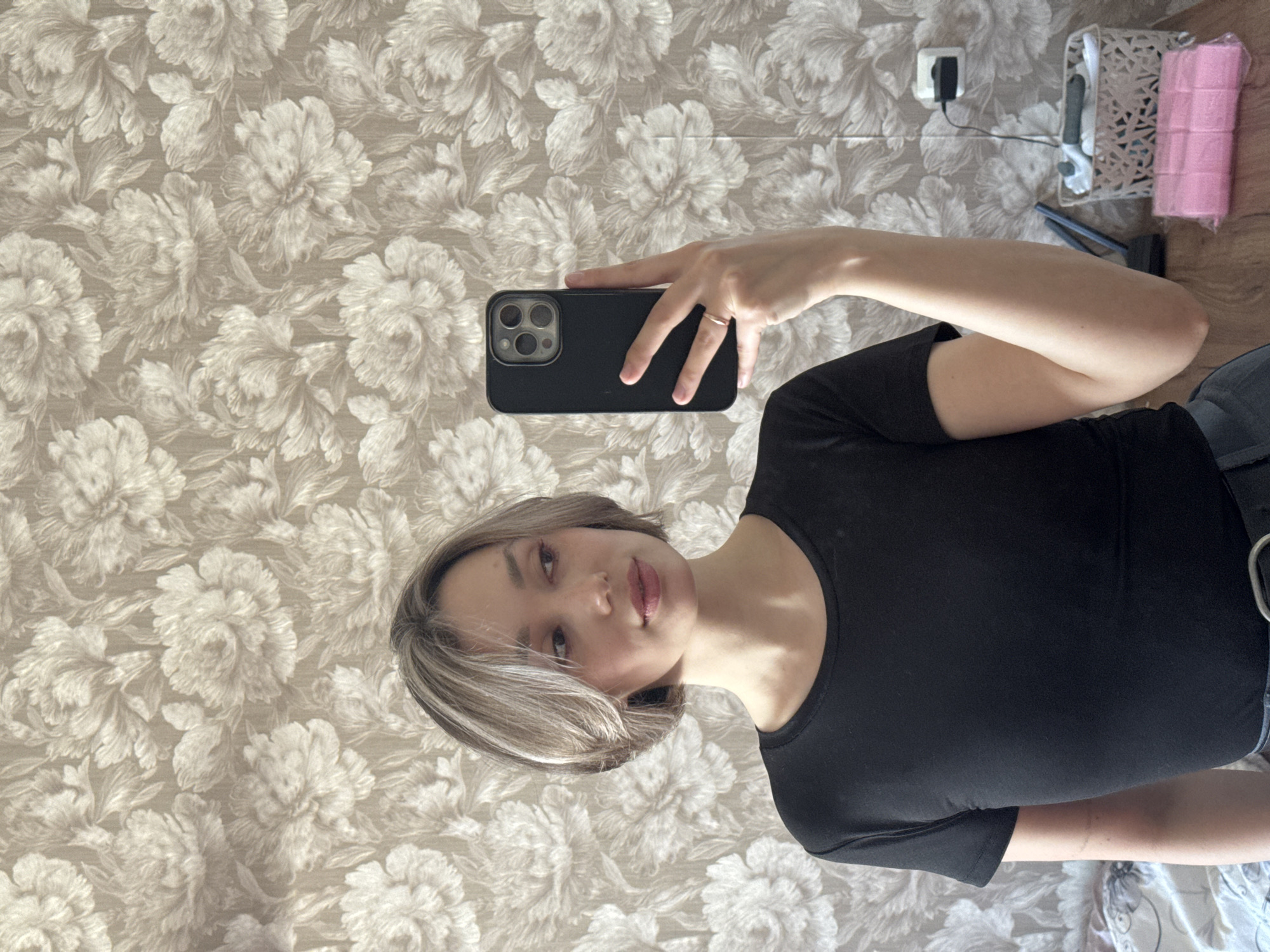 A young woman is holding up a cell phone, likely taking a selfie. She has fair skin and short blonde hair. She is wearing a black short-sleeved shirt. The background features a wallpaper with a repeating floral pattern. There are a few objects visible on a shelf in the background, including what appears to be a white box, a hair straightener, and a roll of pink material. The location is Unknown, Unknown.