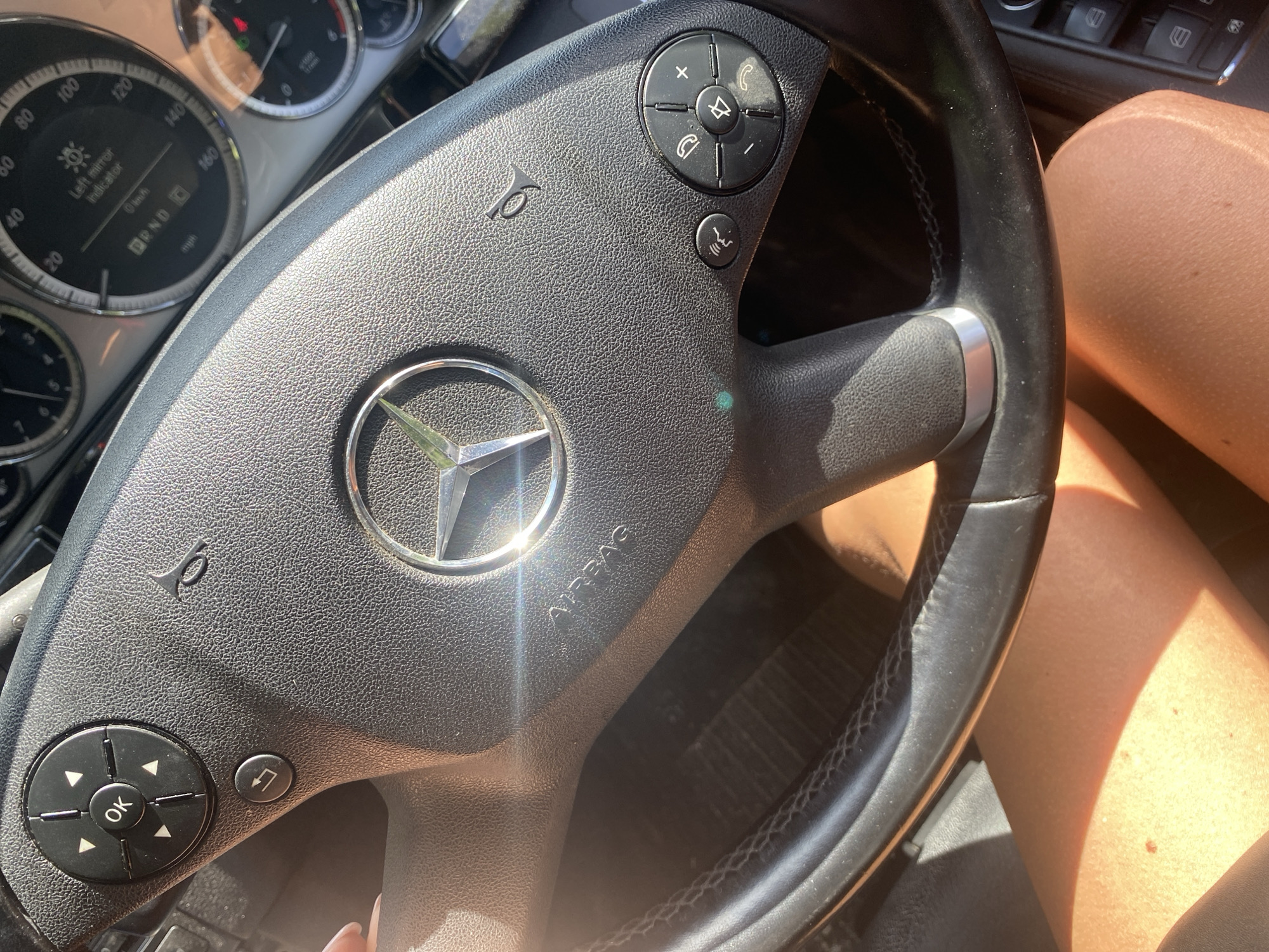 The image provides a close-up view inside a car, focusing on the steering wheel and a portion of the dashboard. The steering wheel, made of black textured material, prominently displays the silver Mercedes-Benz star logo in its center, with "AIRBAG" embossed below it.
Control buttons are visible on both spokes of the steering wheel. On the left, there's a circular pad with an "OK" button surrounded by directional arrows, next to a button featuring a return arrow icon. On the right, another circular pad includes controls for phone calls (answer, hang up), volume adjustment, and mute, accompanied by a voice control button below it. A horn icon is visible on the steering wheel's spokes.
Behind the steering wheel, the instrument cluster is partially visible. The speedometer shows markings up to '160 mph', and a digital display reads "0 km/h", "P R N D" (Park, Reverse, Neutral, Drive), and a message stating "Left mirror indicator".
Bright sunlight streams into the car, casting highlights on the steering wheel and the driver's bare legs, which are visible on the right side of the frame, suggesting a daytime setting. The interior appears to be that of a modern vehicle. This scene is located in Bankya, Bulgaria.