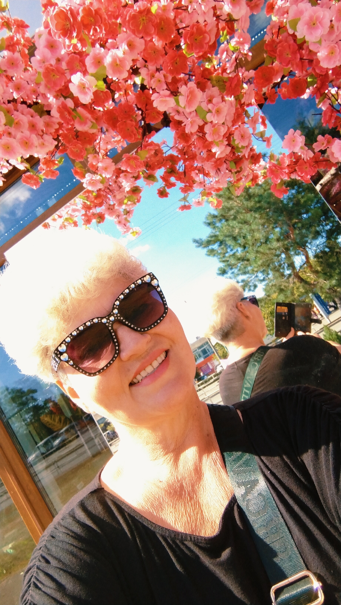 An outdoor, eye-level, medium-shot selfie taken by a woman with blonde hair and sunglasses, smiling at the camera. She is wearing a black shirt and a green strap with text is visible across her shoulder. The woman is positioned in the foreground, with cherry blossoms hanging above and to the left of her. In the background, a reflection in a mirrored surface shows a tree and a blue sky, as well as another person holding a phone. The scene appears to be in Horishni Plavni, Ukraine, near a building with large windows.
