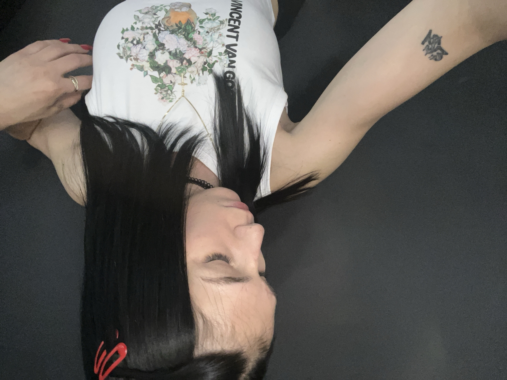 An image shows a person, likely female, with long, straight black hair, lying on a dark, plain surface. The person's head is turned to the side, away from the viewer, with eyes closed or looking downwards, suggesting rest or relaxation.
She is wearing a white sleeveless top with a prominent colorful floral print featuring a vase or pot at the top. The words "VINCENT VAN GOGH" are printed vertically in black capital letters on the right side of the floral design. A gold-colored necklace with a small cross pendant is visible, appearing to be part of a body chain that extends across her torso. A black beaded choker or necklace is also around her neck.
Her left hand, with red painted fingernails and a gold-colored ring on her ring finger, is raised towards her head. Her right arm is extended, revealing a dark tattoo of an East Asian character on her upper arm. Red hair clips or accessories are visible in her black hair near the bottom of the frame. The background is a uniform dark color, offering no specific location details.