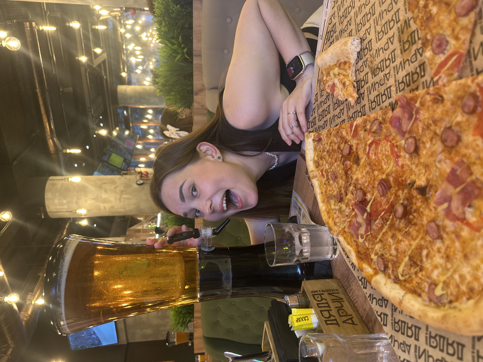 A woman with dark hair and green earrings poses with a large pizza and beer tower at the DADYA Archi Beer Bistro in Perm, Russia. She is smiling with her mouth open, and is wearing a smartwatch. The pizza is on cardboard marked with the bistro's name. There are several glasses on the table. The background shows the bistro's interior with warm lighting.