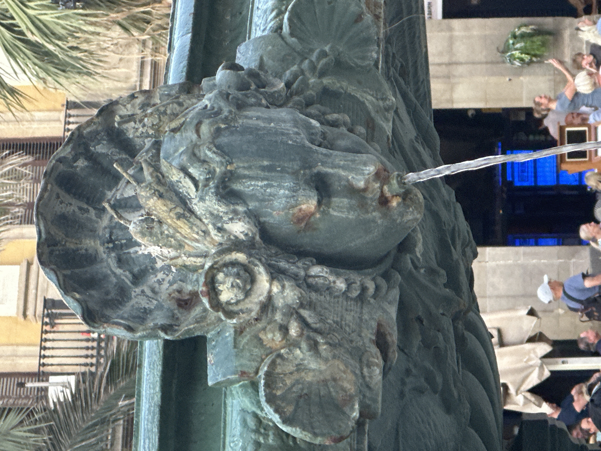 The image shows a close-up view of a decorative gargoyle on a fountain, likely in a public square in Barcelona, Spain. The gargoyle is sculpted in the form of a human face, with a stylized headdress resembling a fan or shell. Water is spouting from its mouth, creating a thin stream. The gargoyle is made of a weathered, greenish-blue metal, possibly bronze, with visible signs of rust and patina. In the background, palm fronds are visible, along with architectural elements of a building with a balcony and shutters. The lower portion of the image shows a blurred scene of people, suggesting the fountain is located in an area with public seating or a gathering space. One person in the foreground is wearing a blue shirt and a white cap, and another is holding a smartphone. The overall impression is one of a historic urban setting.