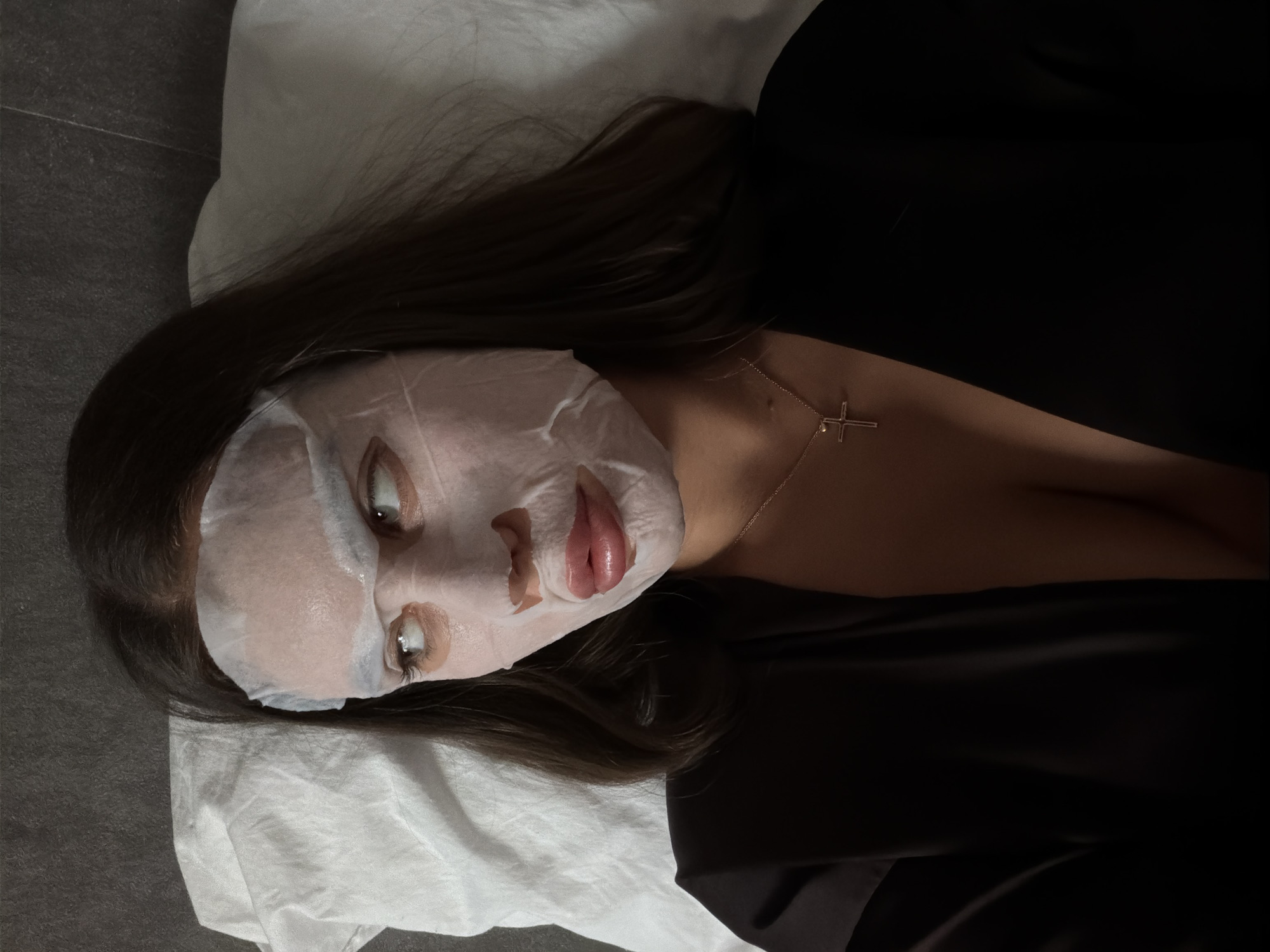 A woman with dark hair is lying on a white pillow, wearing a sheet mask on her face. She is also wearing a gold necklace with a cross pendant. The background is a dark gray surface, possibly a floor or wall. There is no evidence in the image to suggest that the location is Krasnoyarsk, Russia.