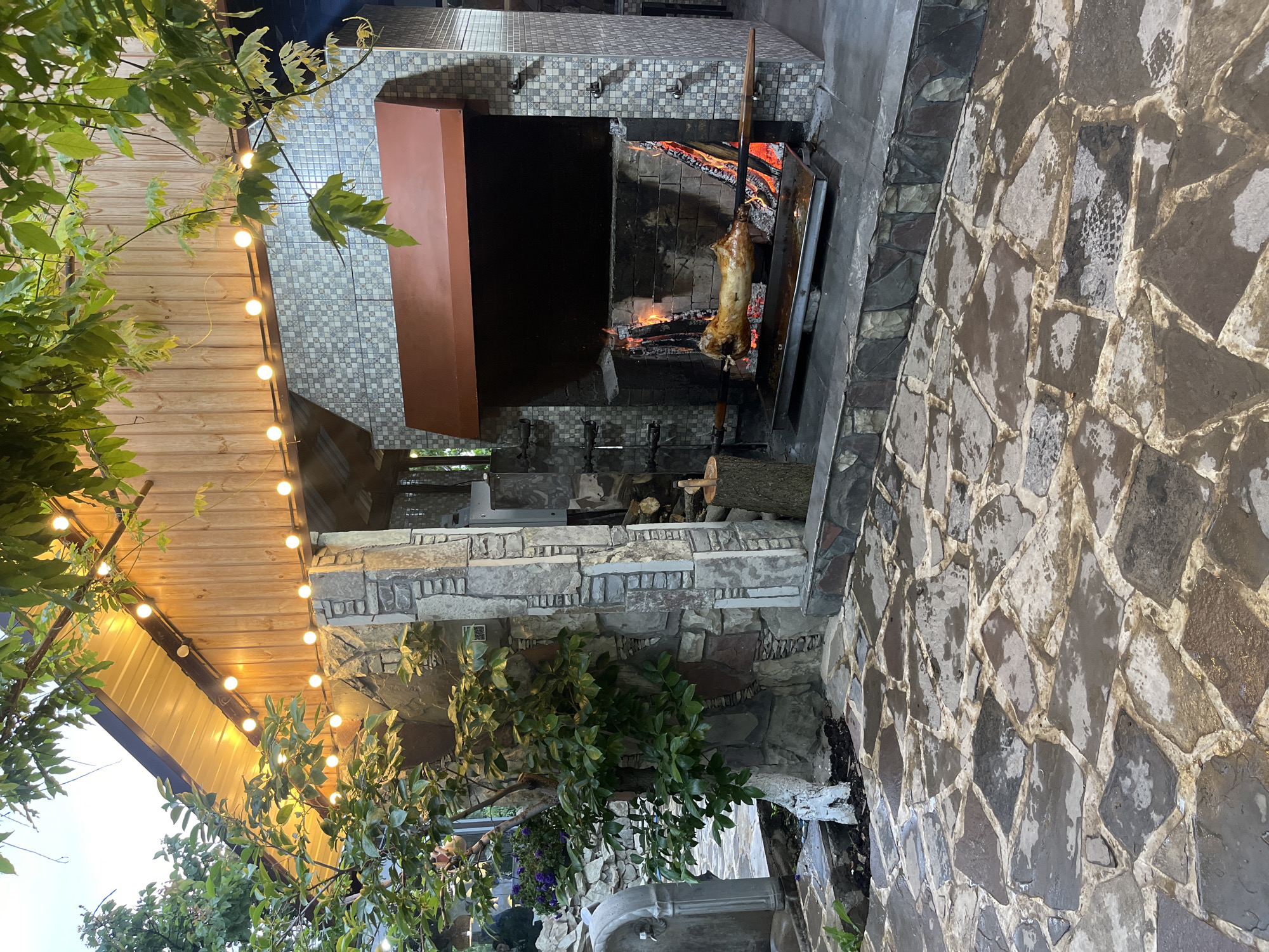 The image shows an outdoor cooking area in what appears to be a patio or garden setting. A whole animal, likely a pig, is being roasted on a spit over an open fire. The cooking area is constructed of stone and brick. There is a canopy above with string lights hanging from it. The ground is paved with stone tiles. Plants and trees are visible around the cooking area. The image does not contain evidence to confirm that it is taken in Miatli, Russia.
