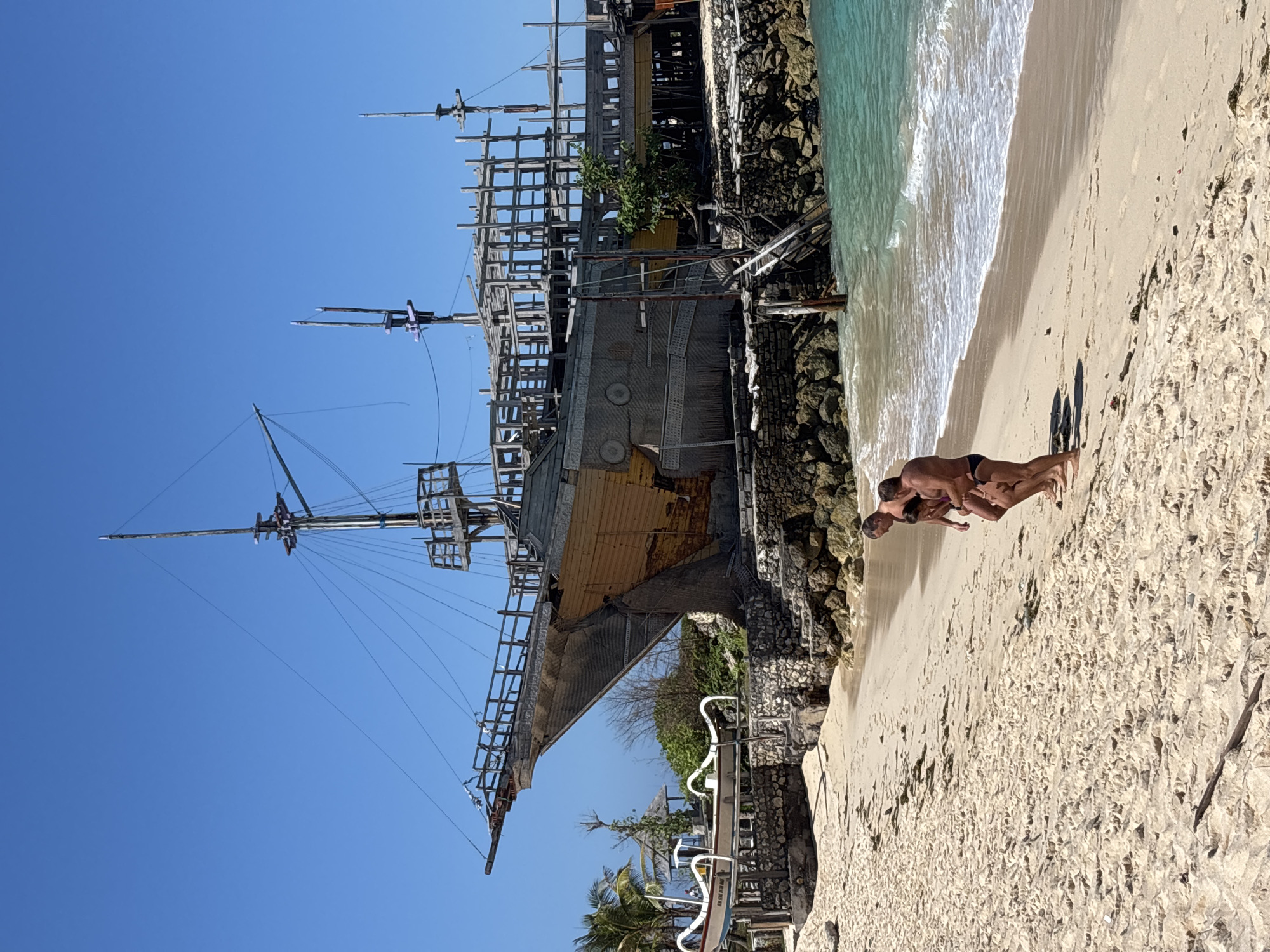 A partially constructed wooden pirate ship is prominently displayed on a rocky shoreline adjacent to a sandy beach in Kuta Selatan, Indonesia. The ship features a weathered yellow hull and a skeletal wooden superstructure. Two men and one woman, all in swimwear, walk along the shoreline in the foreground, with the ocean lapping at the beach. The water is a clear turquoise, and the sky is a vibrant blue. Several smaller traditional Balinese boats are visible on the shore near the ship. The scene suggests a tropical beach location with a unique architectural feature.